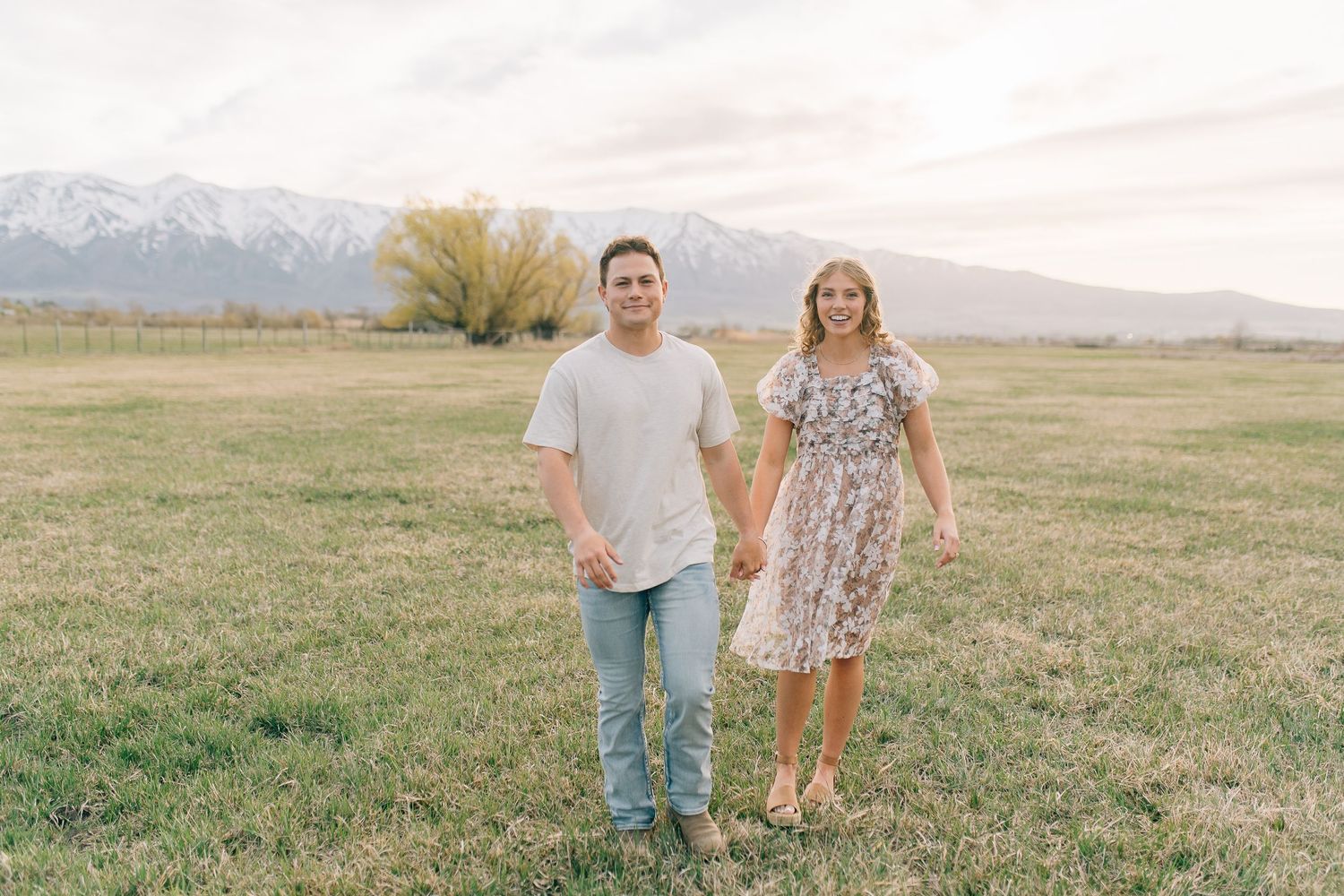 Two people stand hand-in-hand in an open field with mountains and a cloudy sky in the background.