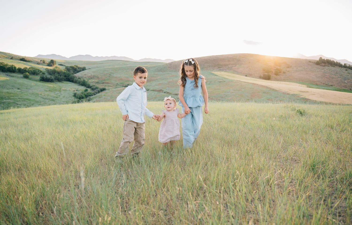Three children holding hands in a sunlit green meadow with rolling hills at golden hour.