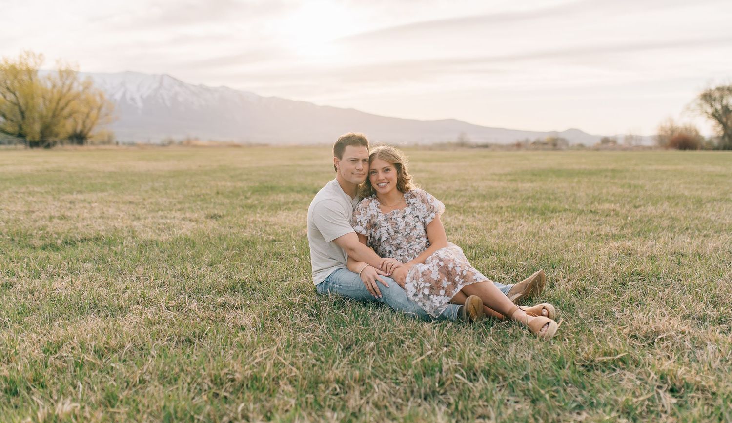 A couple sits together on grassy field with mountains visible in the background during golden hour.