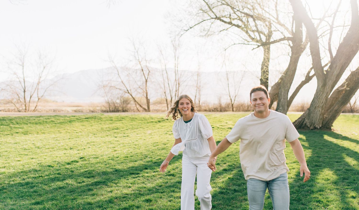 People joyfully running together on a grassy field with mountains and bare trees in the background.