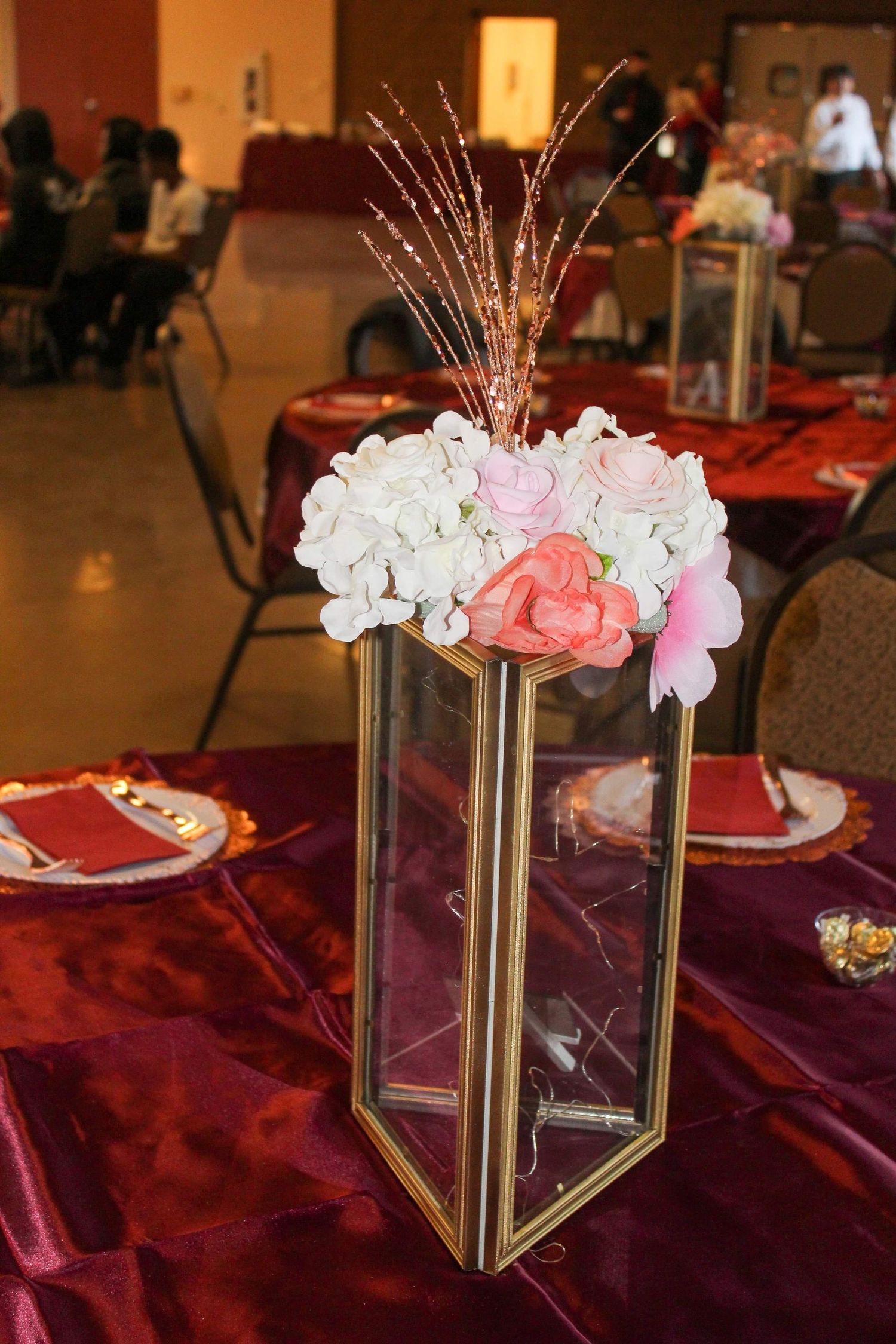A modern geometric gold and glass vase filled with pink and white hydrangeas on a burgundy tablecloth at an event setting.