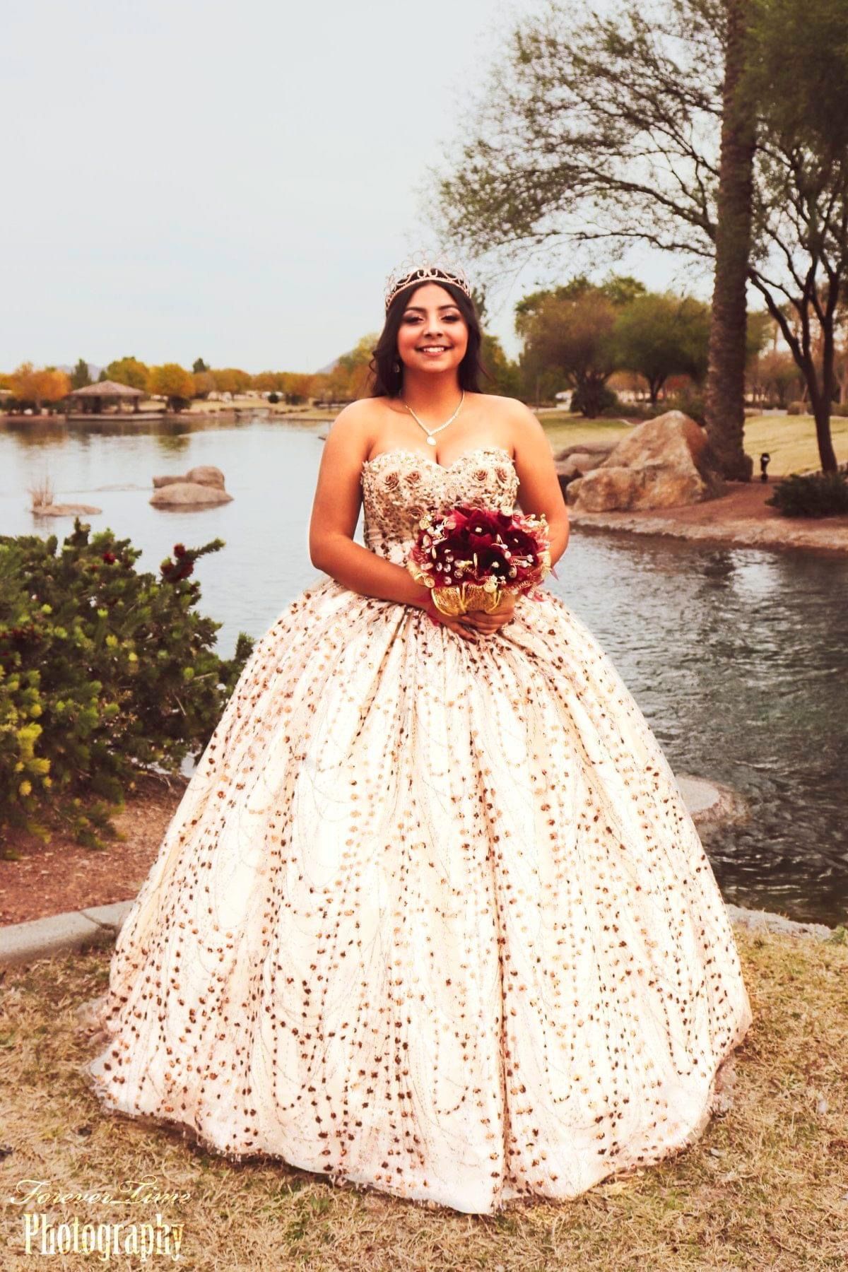 A person in a white and gold formal ball gown with sequins holds a purple bouquet by a waterfront landscape. for a quinceanera .
