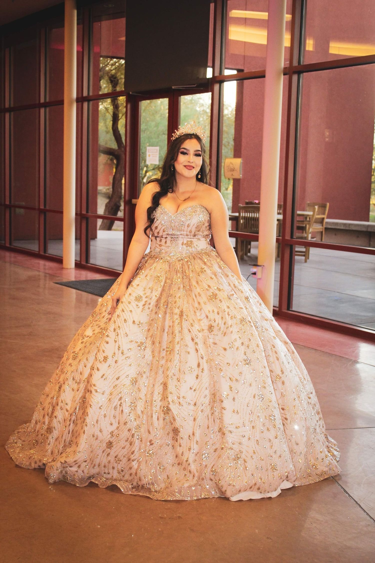 A person poses in a sparkly champagne-colored ball gown with a full skirt and strapless bodice outside a building.