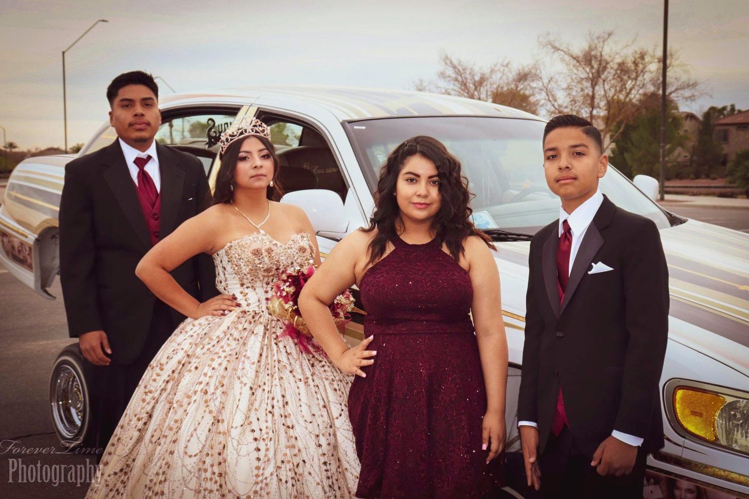 Four elegantly dressed prom guests pose in formal attire next to a white car during sunset.