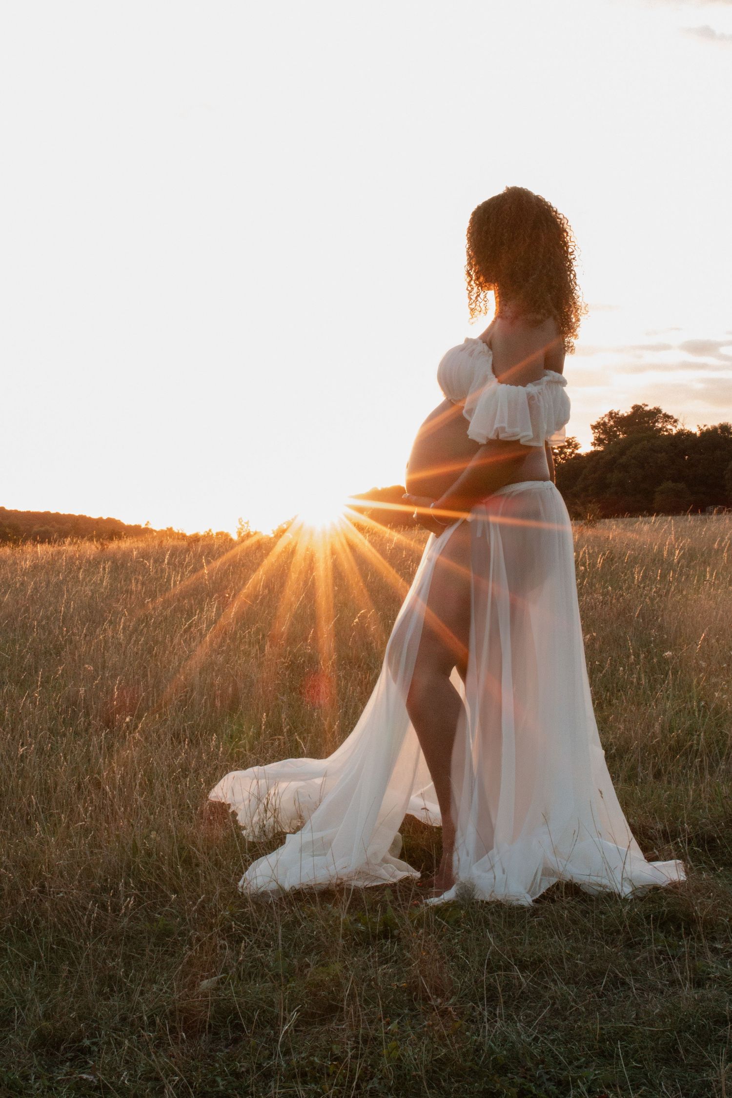 Maternity photograph of Figure in flowing white dress silhouetted against dramatic sunset in meadow.