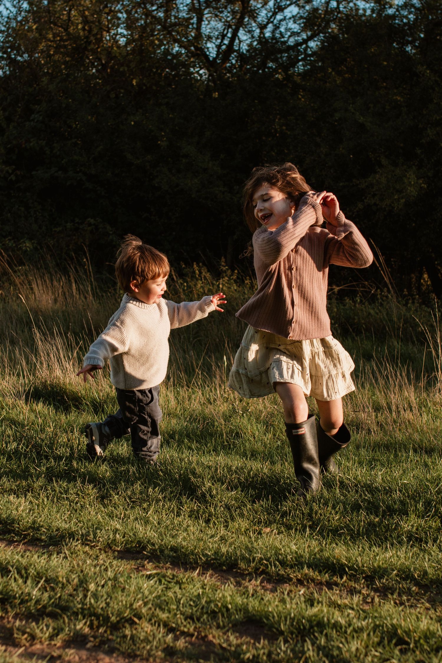 Two young children playing and running together in a golden hour sunlit grassy field with trees in the background.