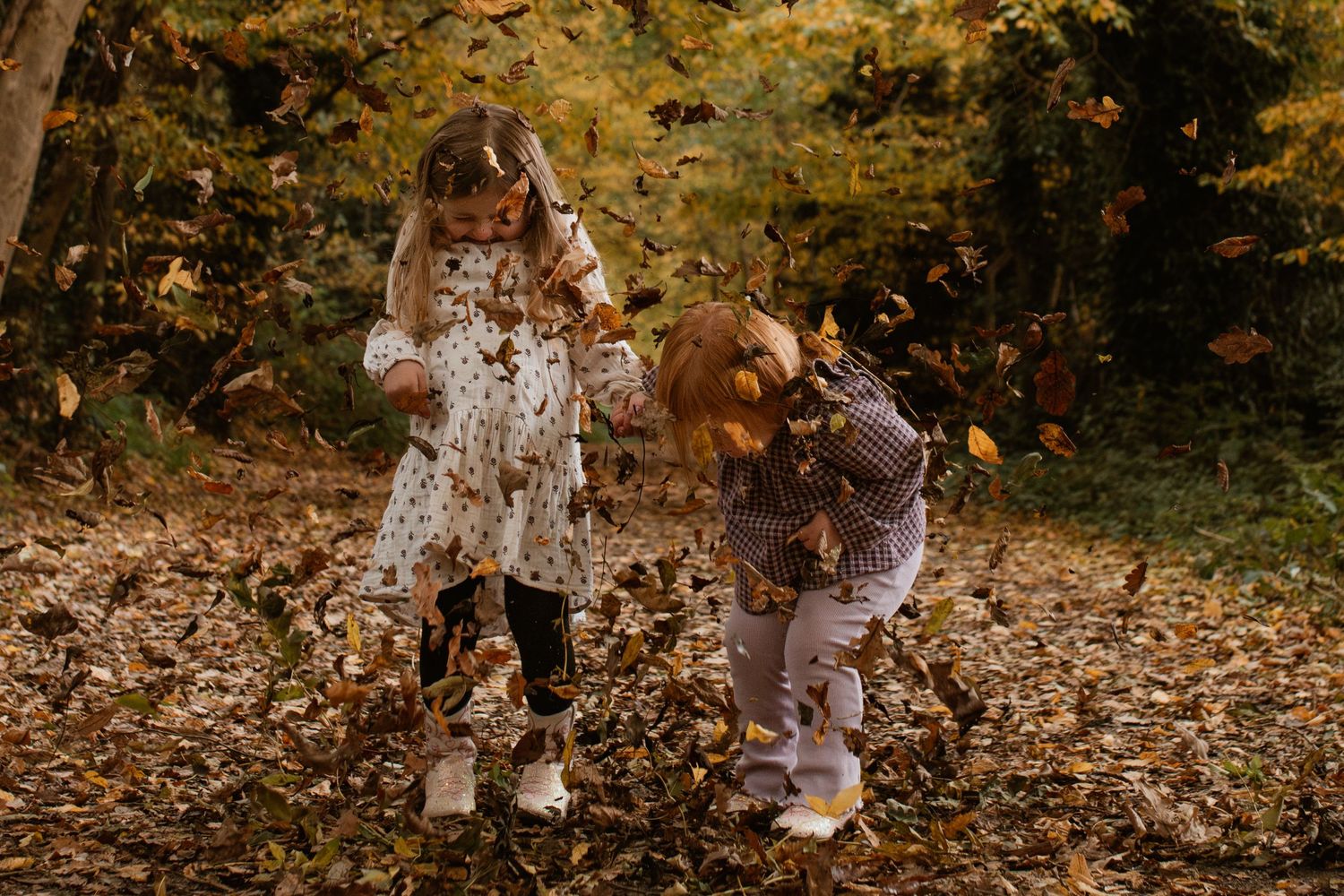Two children playing joyfully in autumn leaves in a forest, surrounded by golden fall foliage.
