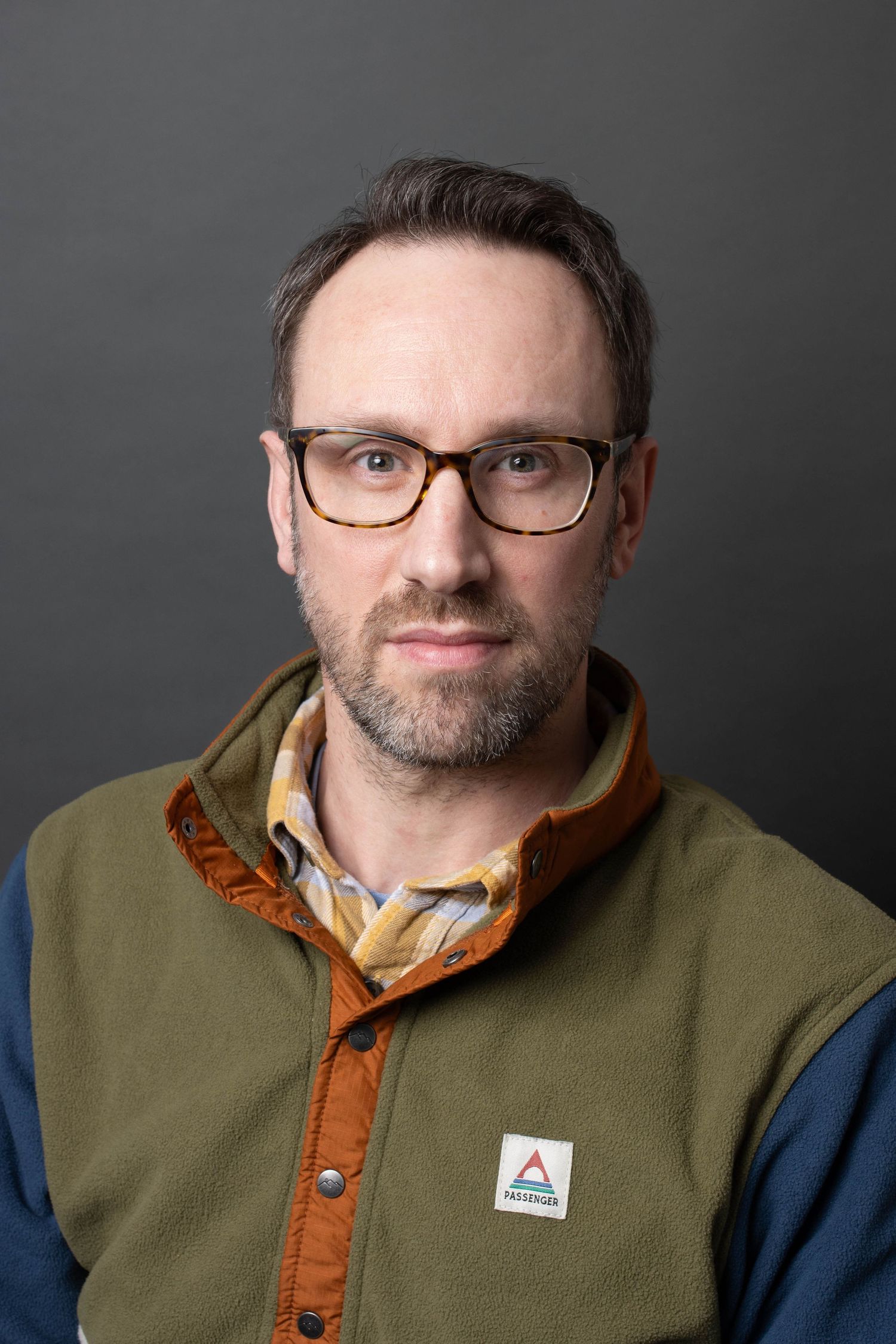 Portrait of person wearing olive green vest and glasses against dark gray background.