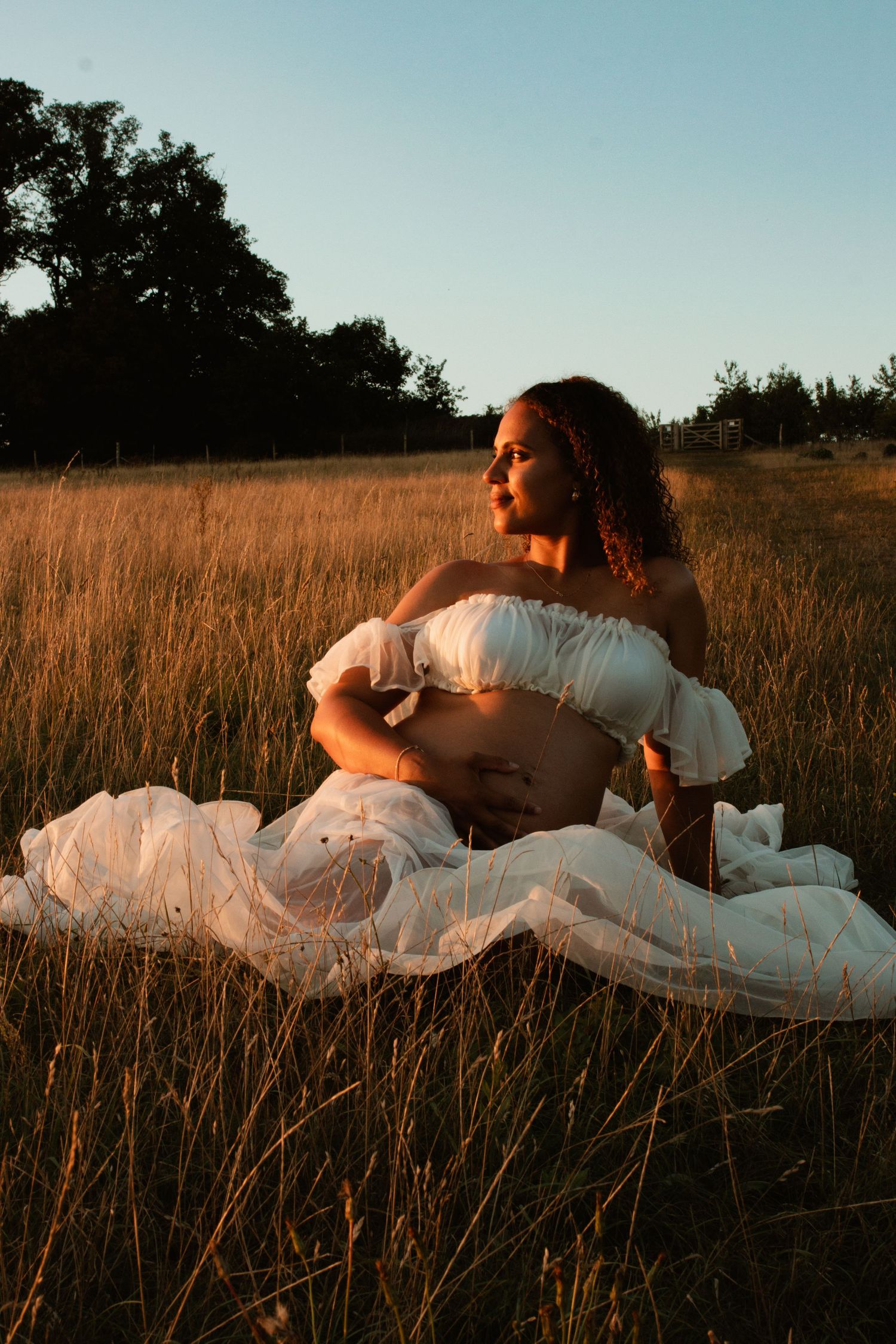 Pregnant woman in white off-shoulder dress sitting in golden field at sunset, cradling her baby bump.