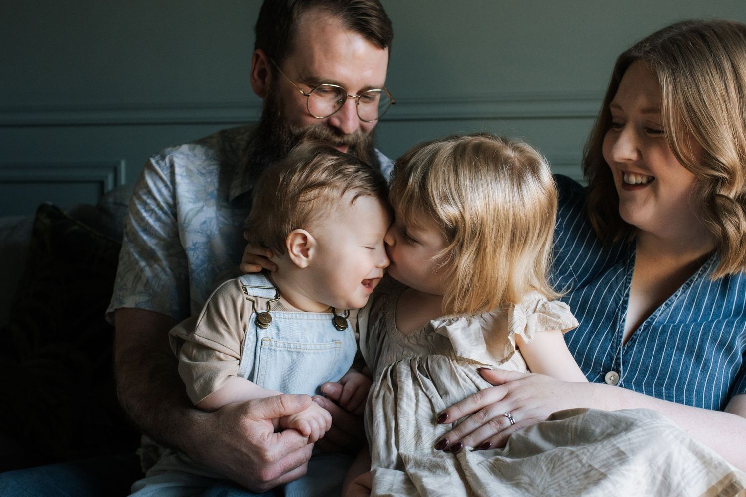 A tender family moment with parents and two young children sharing a sweet kiss indoors.