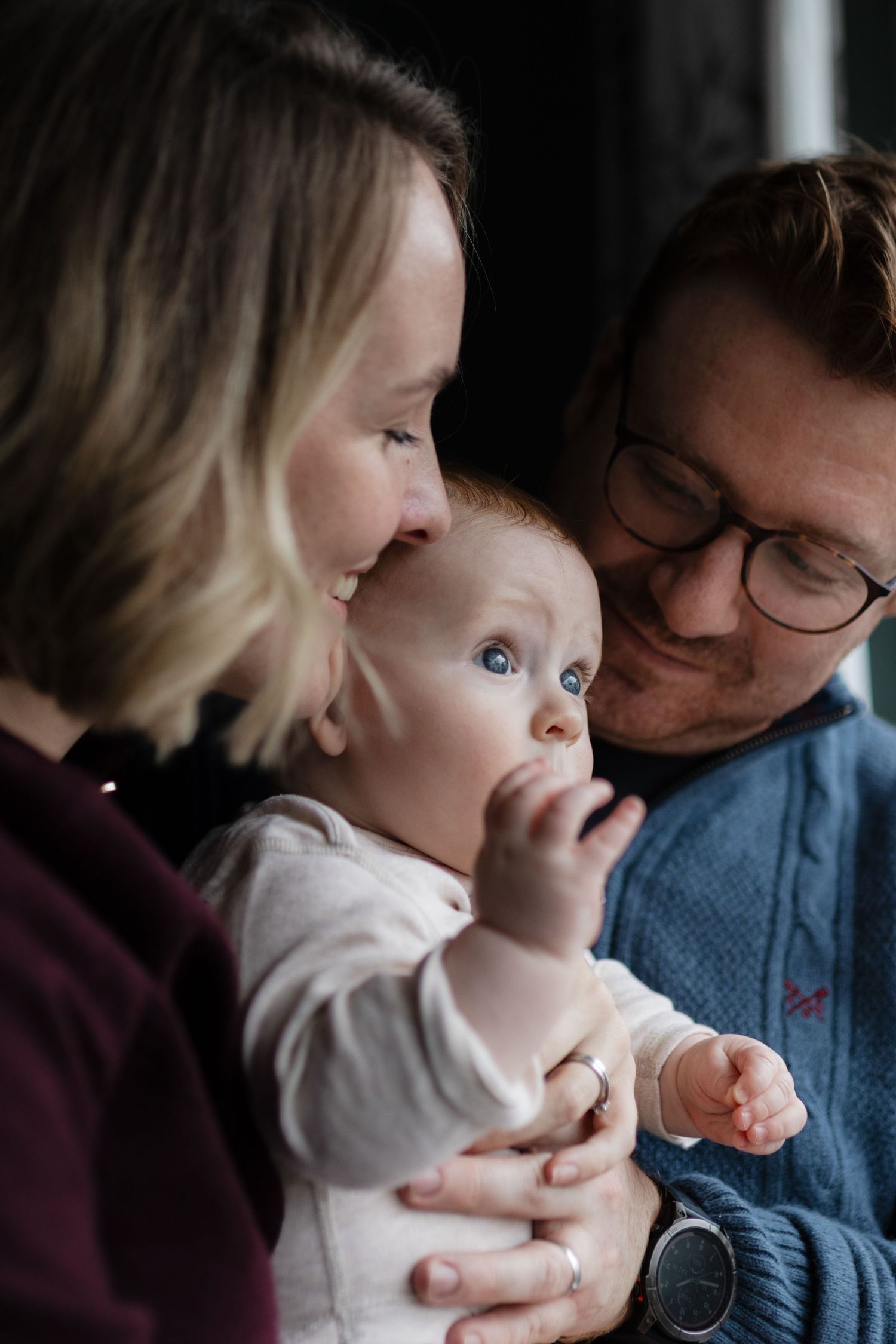 A young family with a mother, father wearing glasses, and a blue-eyed baby sharing a tender moment together.