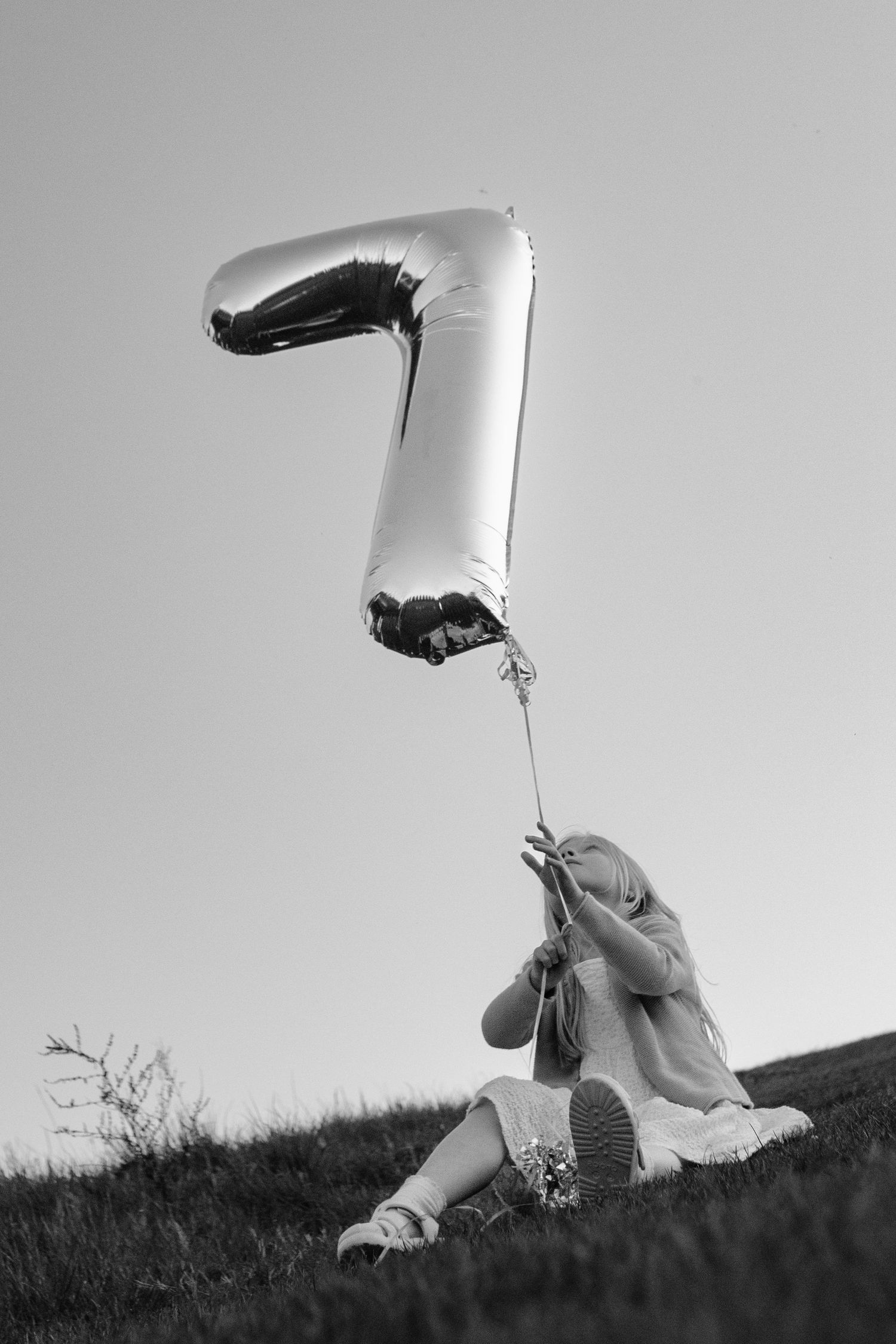 A young girl sits on grass holding a large number 7 mylar balloon outdoors, captured in black and white photography.