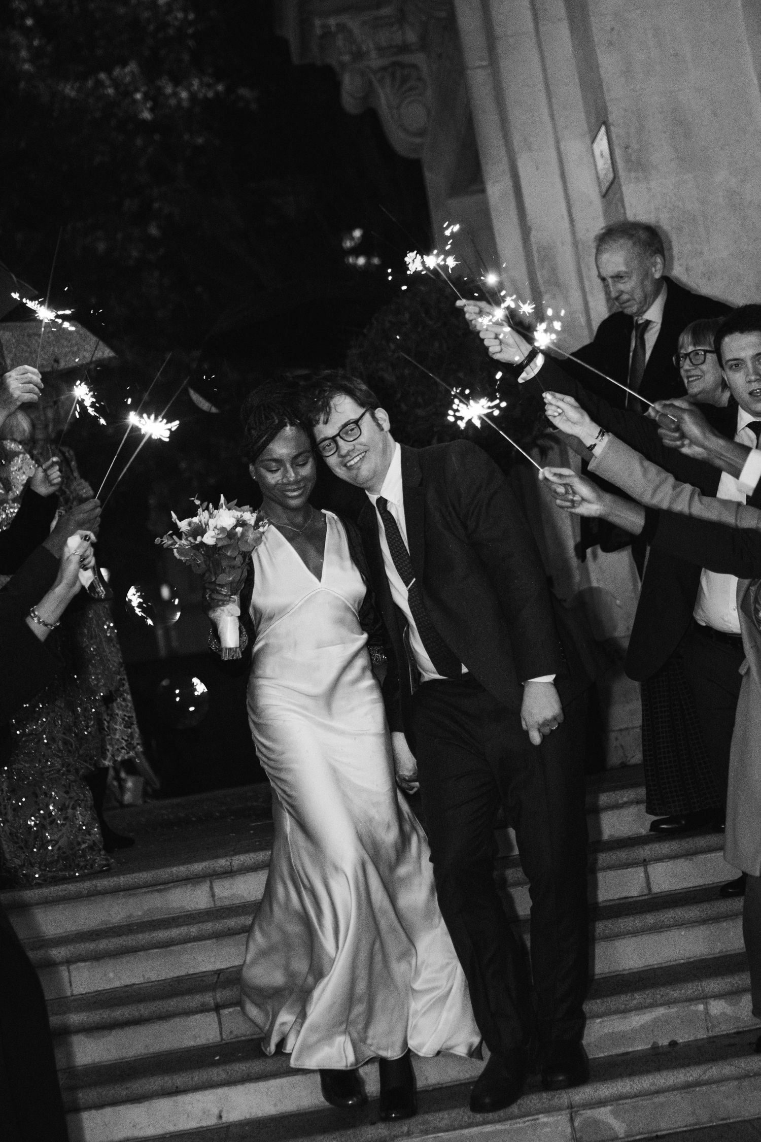 Newlywed couple exits their wedding ceremony down steps while guests wave sparklers in celebration at night.