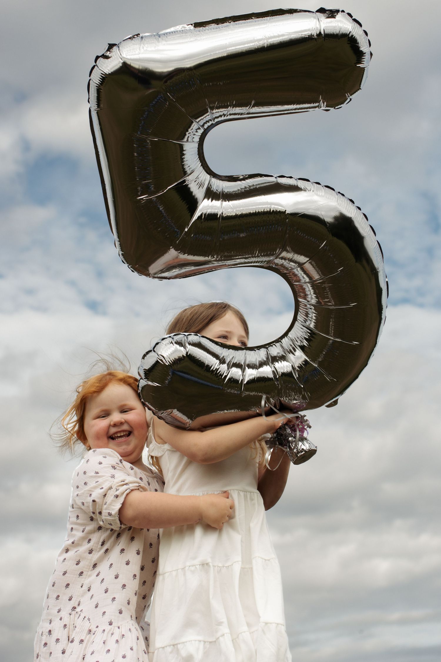Two young girls in white dresses joyfully hold a large silver number 5 balloon against a cloudy sky.