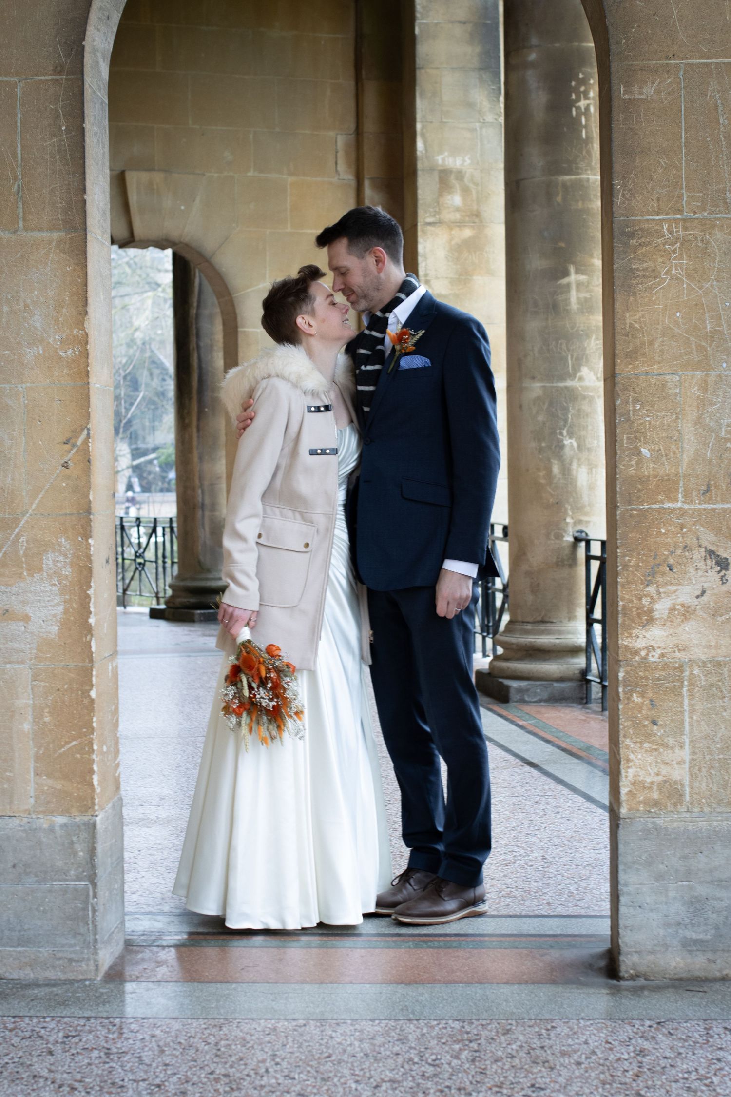A romantic wedding couple shares an intimate moment under a stone archway with the bride holding an autumn bouquet.