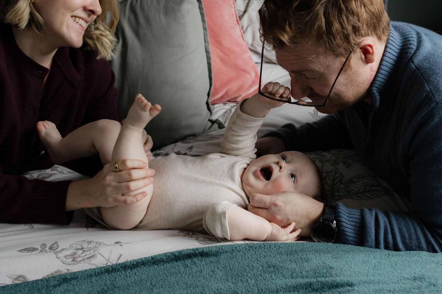 Parents smiling and bonding with their laughing baby lying on a bed with teal blanket and pink pillows.