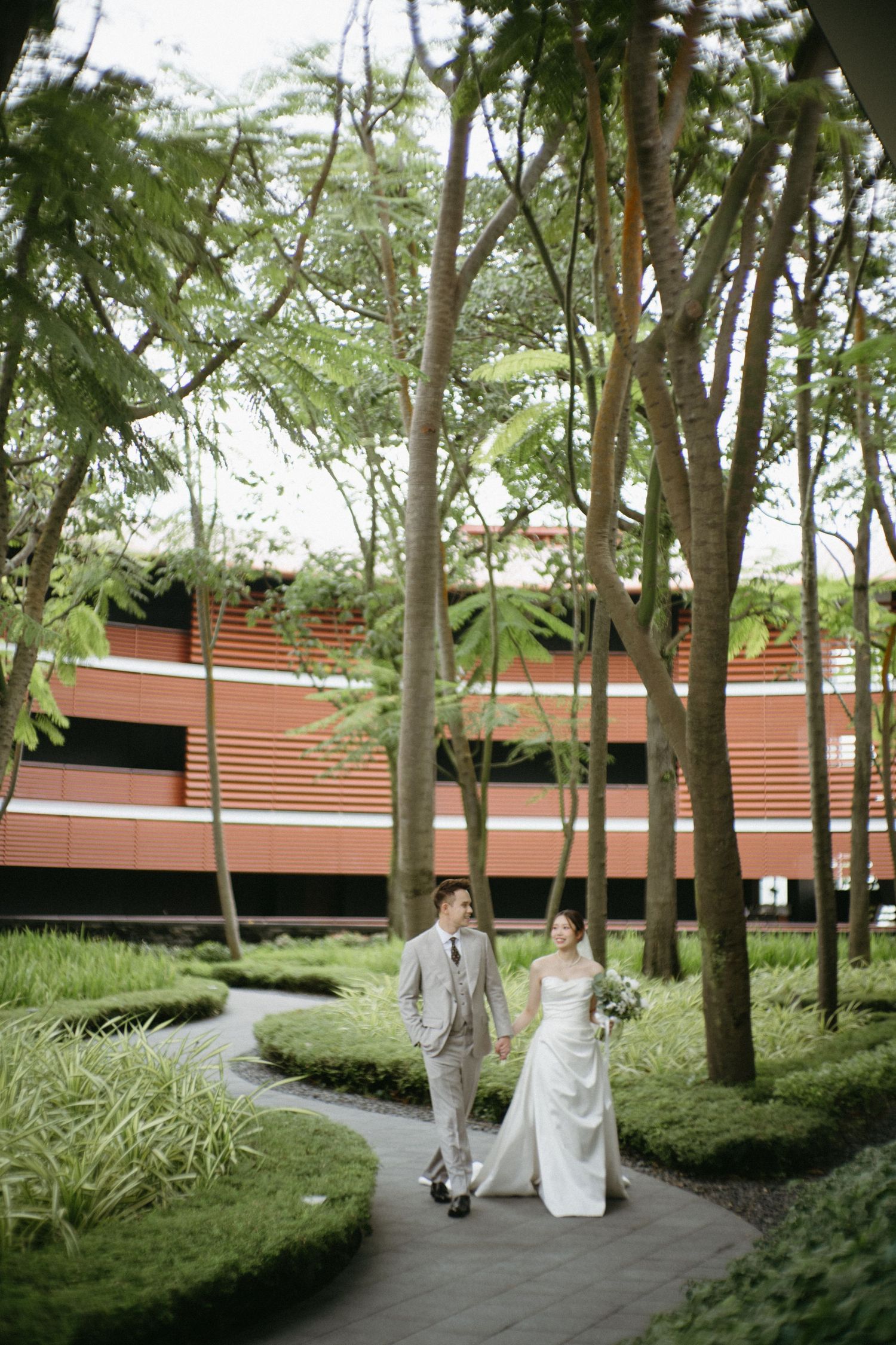 Two people in wedding attire walk along a winding garden path beside a modern building with terra cotta colored walls.