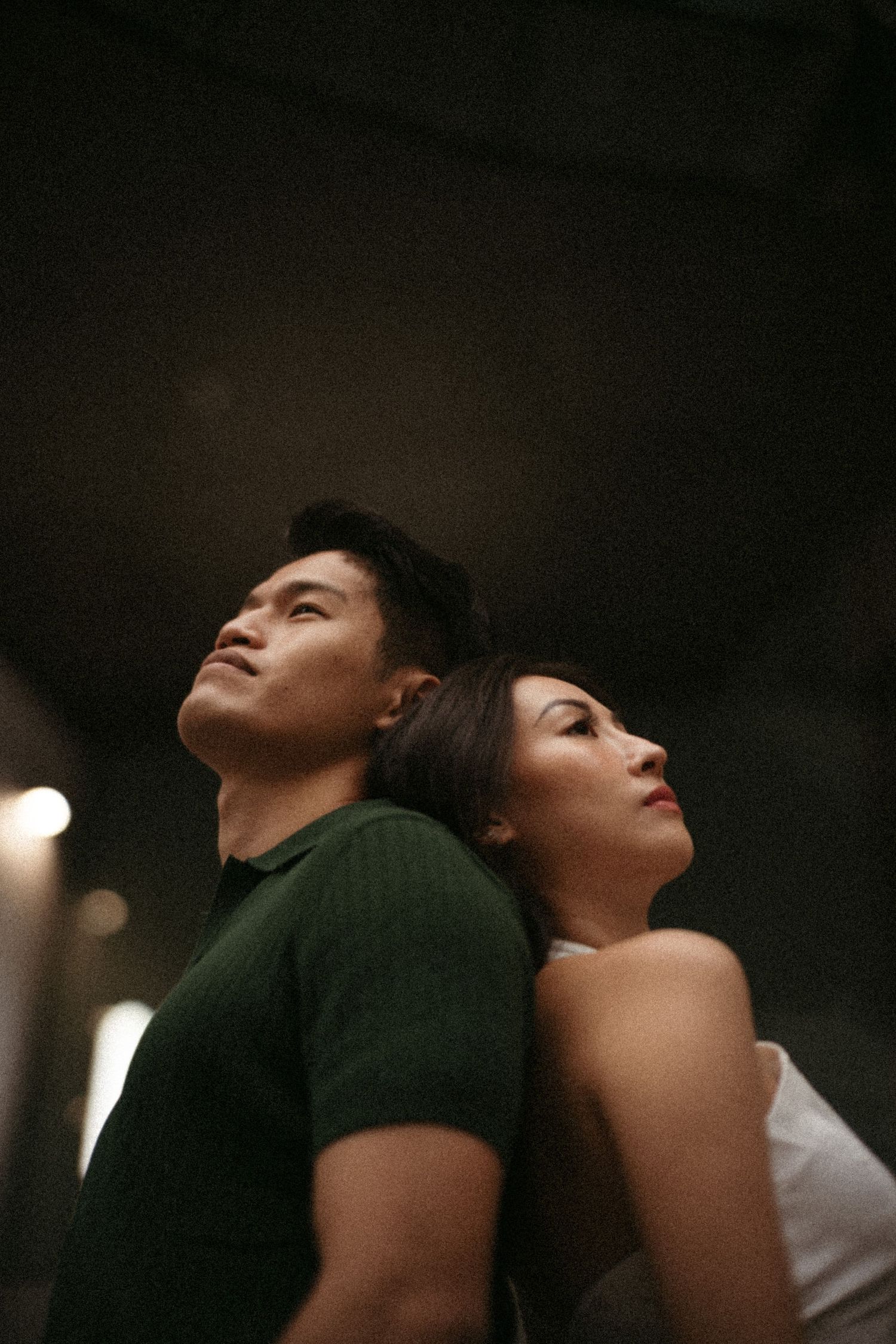 Two people standing back to back in dramatic lighting, with person in green shirt leaning against person in white.