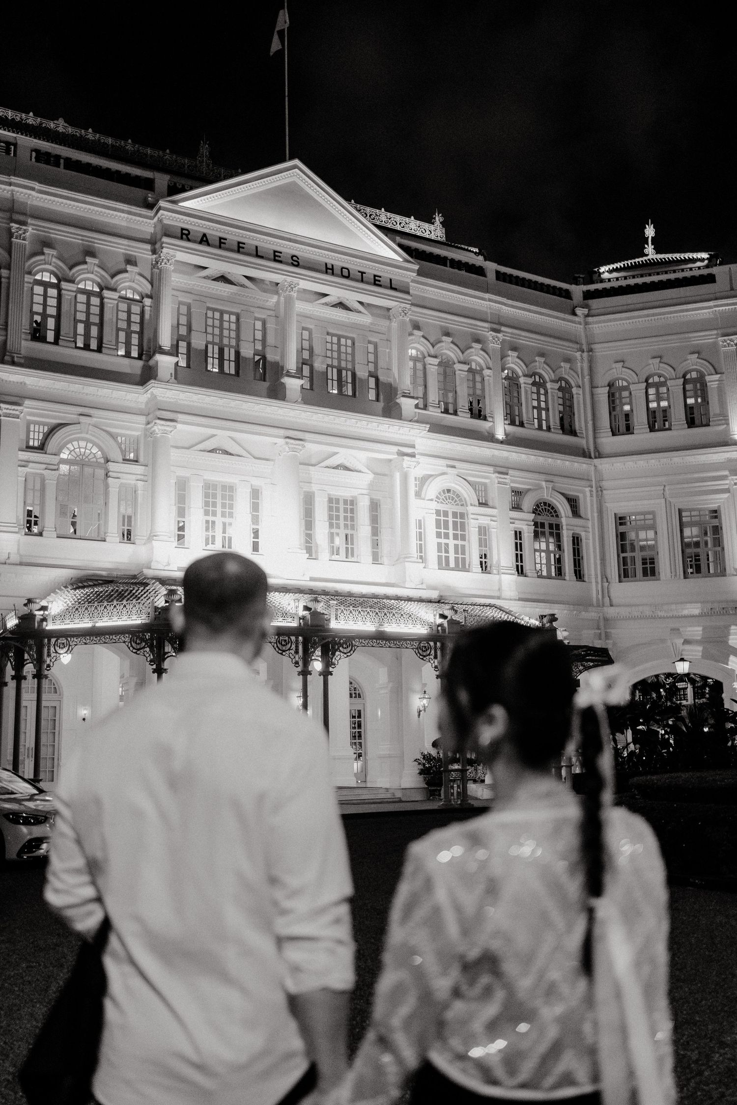 A black and white nighttime view of the illuminated Raffles Hotel facade in Singapore with silhouetted figures in foreground.