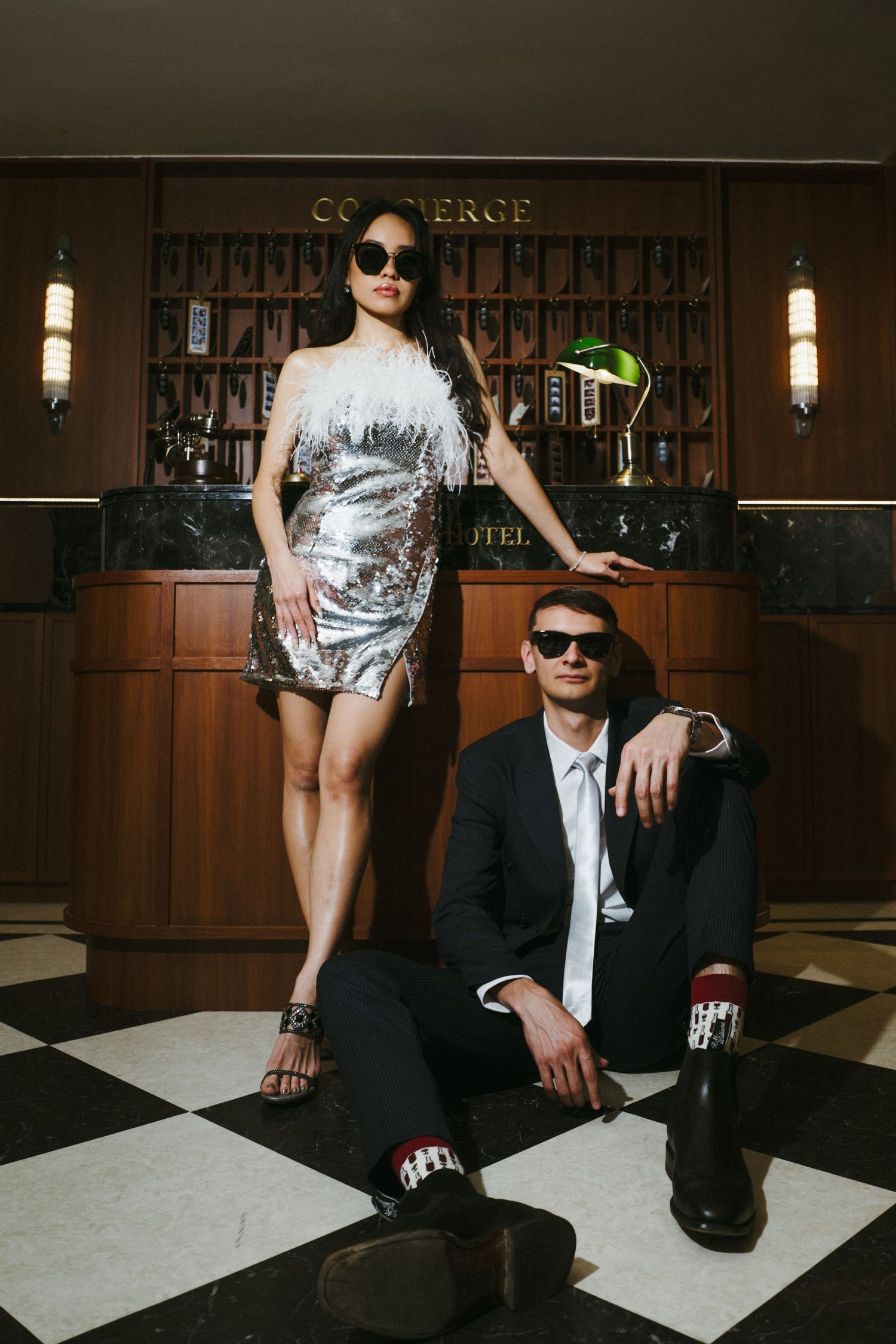 Two people in contrasting outfits pose on a checkered floor in front of a wooden bar counter.