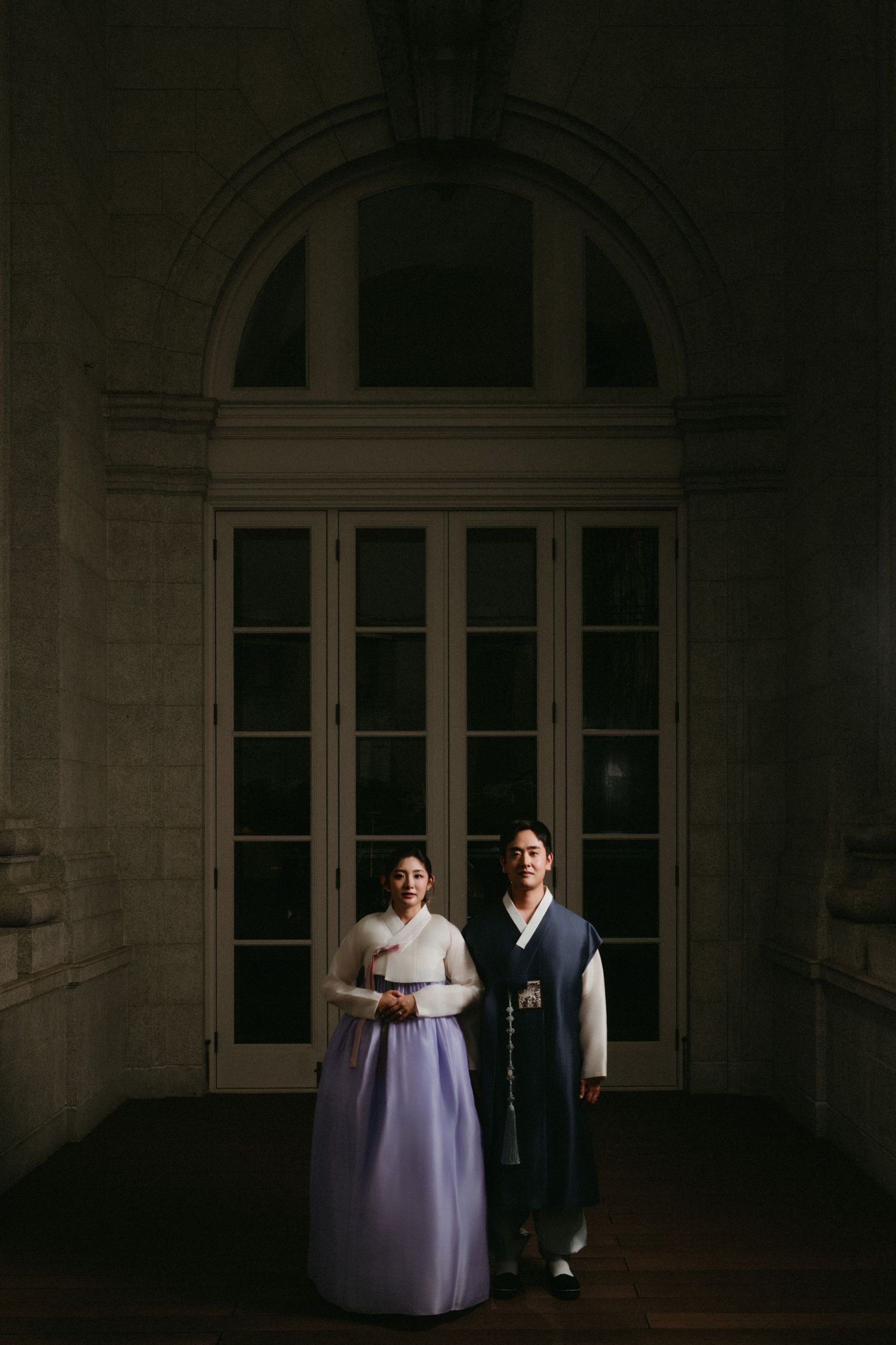 A couple stands in traditional formal attire against a grand doorway with ornate architectural details in dark moody lighting.