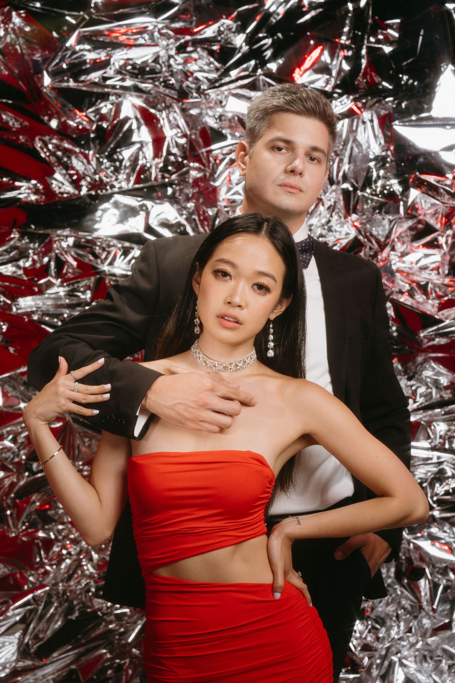 Fashion photo with dramatic silver foil backdrop featuring a couple in red and black formal attire striking a posed embrace.