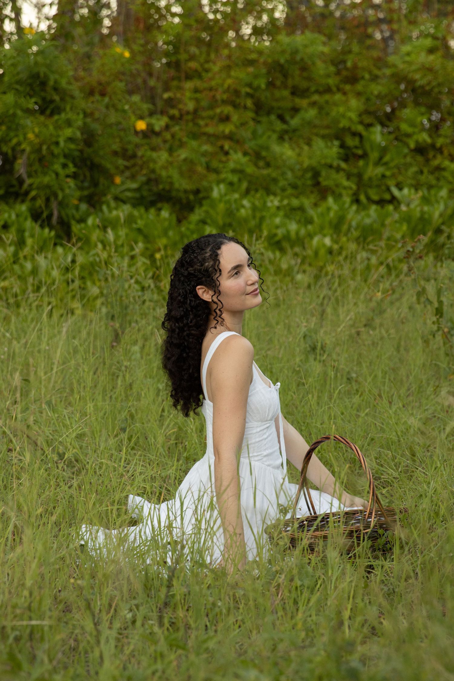 A person in a white dress sits peacefully in tall grass with a woven basket on a summer day.