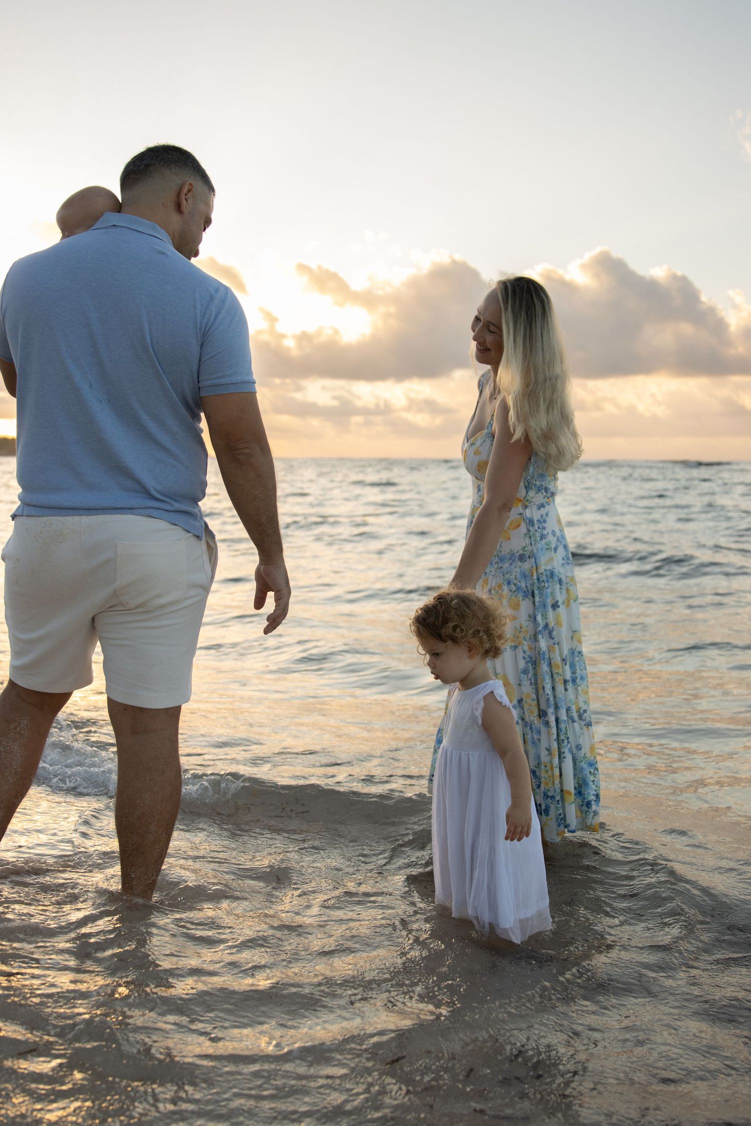 Couple standing in ocean waves during golden sunset beach photoshoot wearing white and blue outfits.