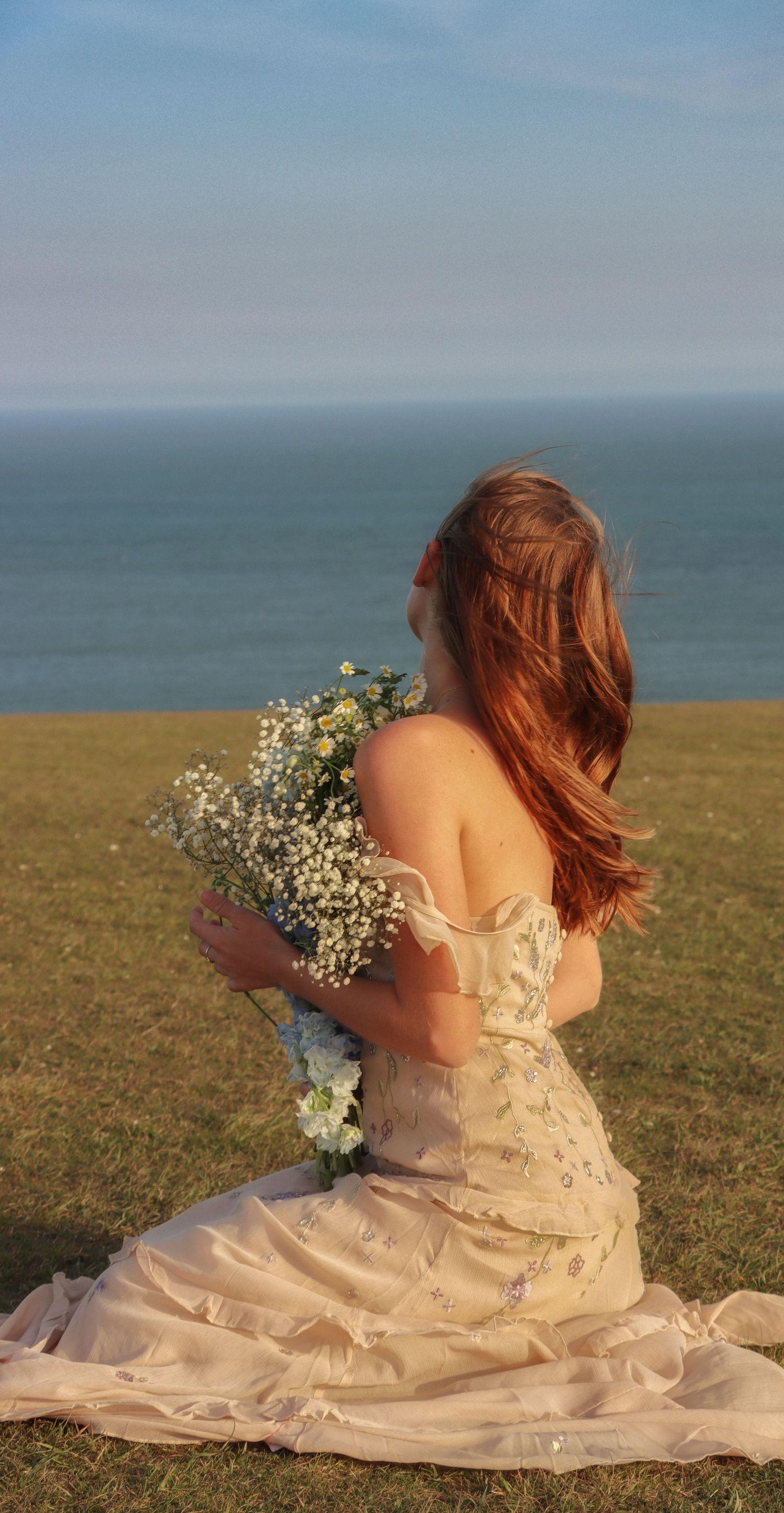 Person in flowing dress sits on coastal cliff holding white wildflowers at sunset. By Chrissie McLean Photography.