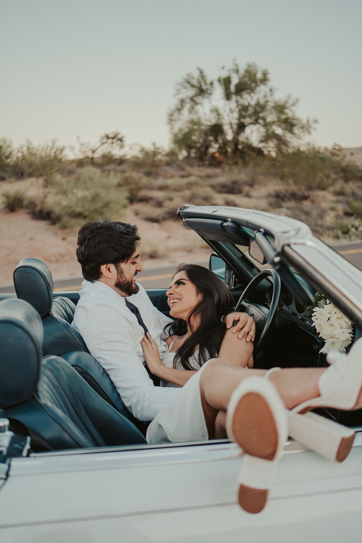 A couple shares intimate moments in a vintage convertible car against a desert backdrop.