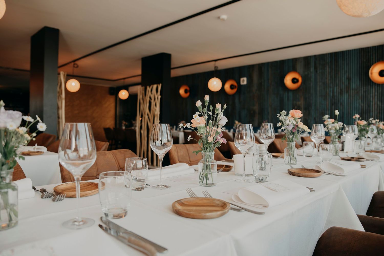 Elegant wedding reception table setting with white linens, wooden chargers, and soft glowing pendant lights overhead.