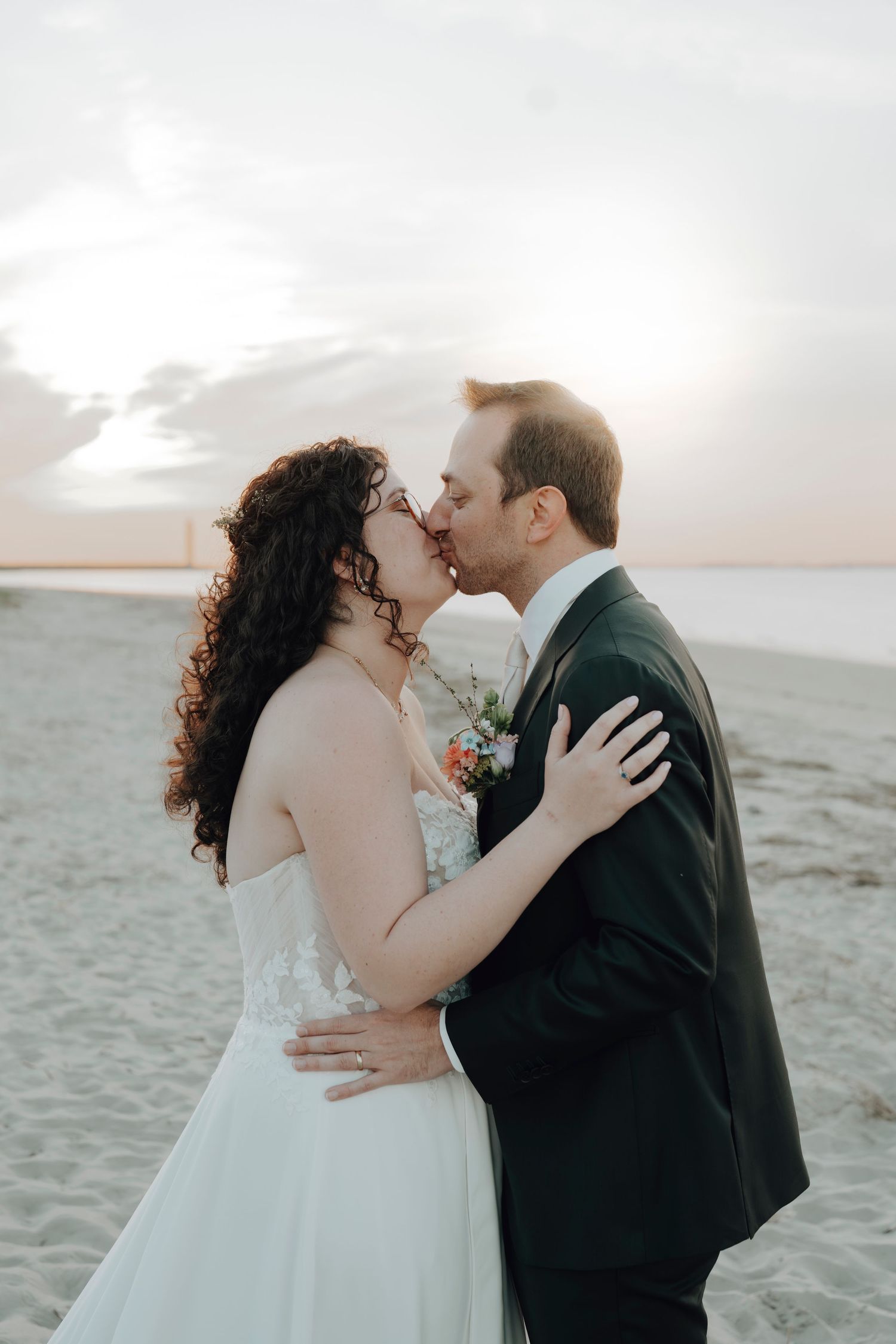 A couple in wedding attire embrace and kiss on a sandy beach at sunset.