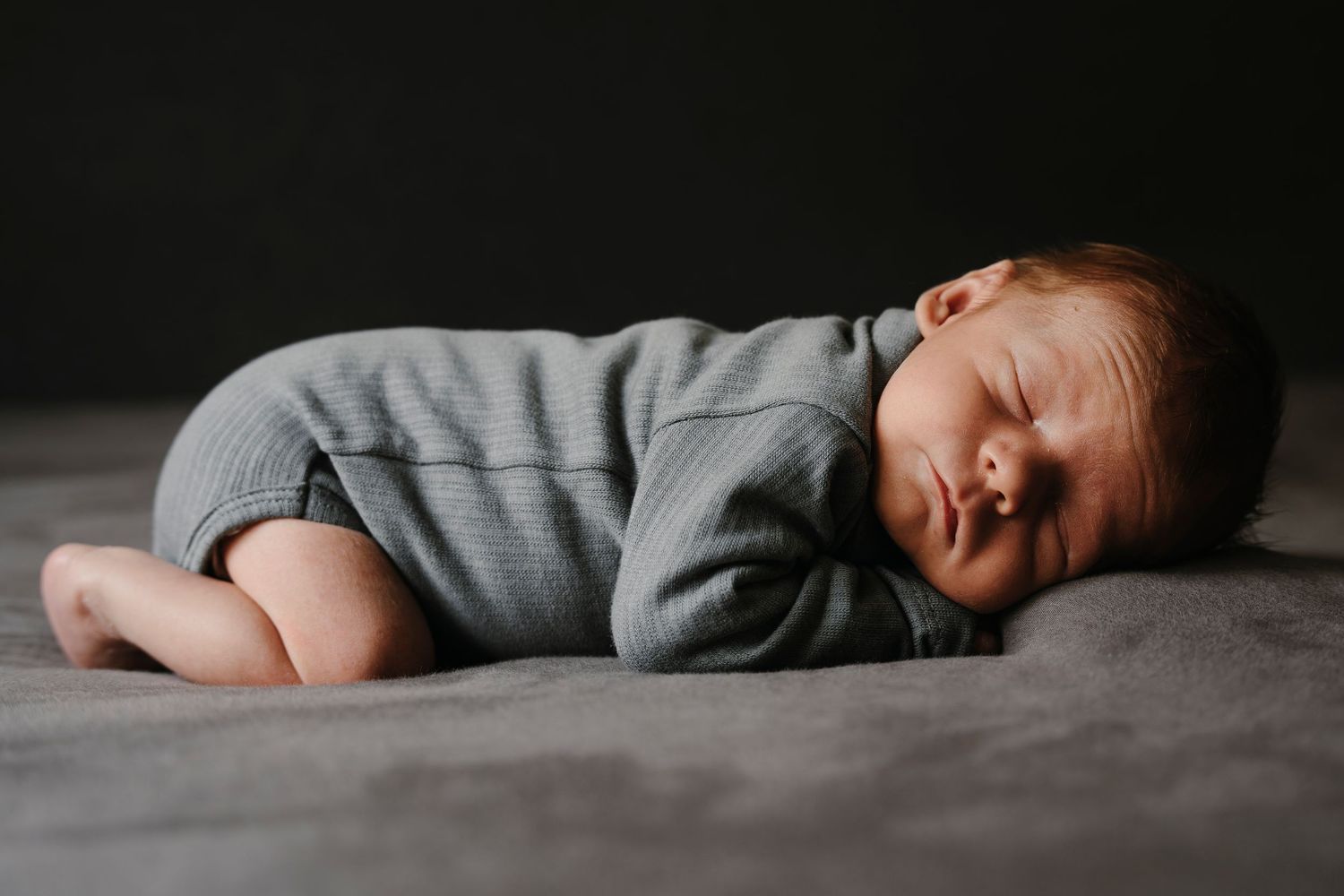 A sleeping baby wearing a gray onesie lies peacefully on a dark gray blanket in dim lighting.
