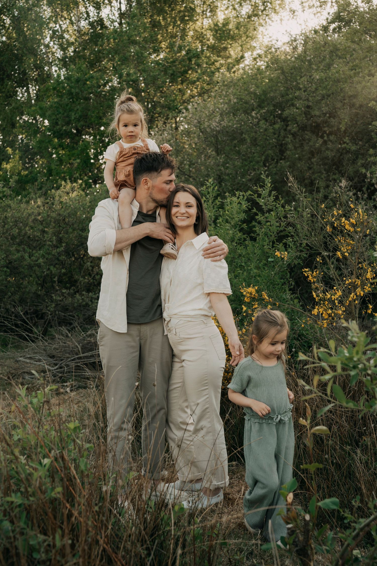 A family poses together outdoors in neutral clothing amid tall grass and greenery at sunset.