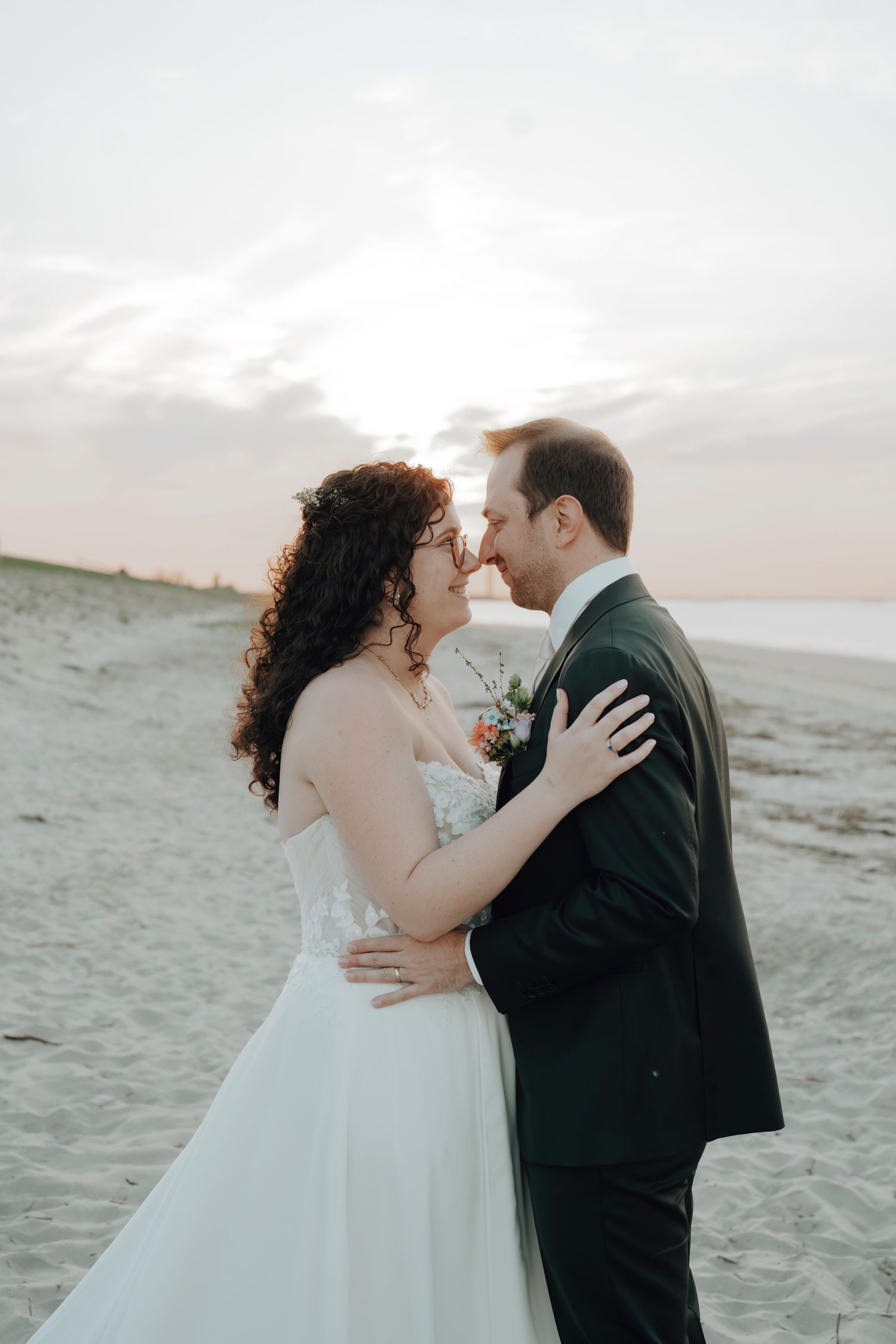 Newlywed couple sharing an intimate moment on a sandy beach at sunset with cloudless sky.