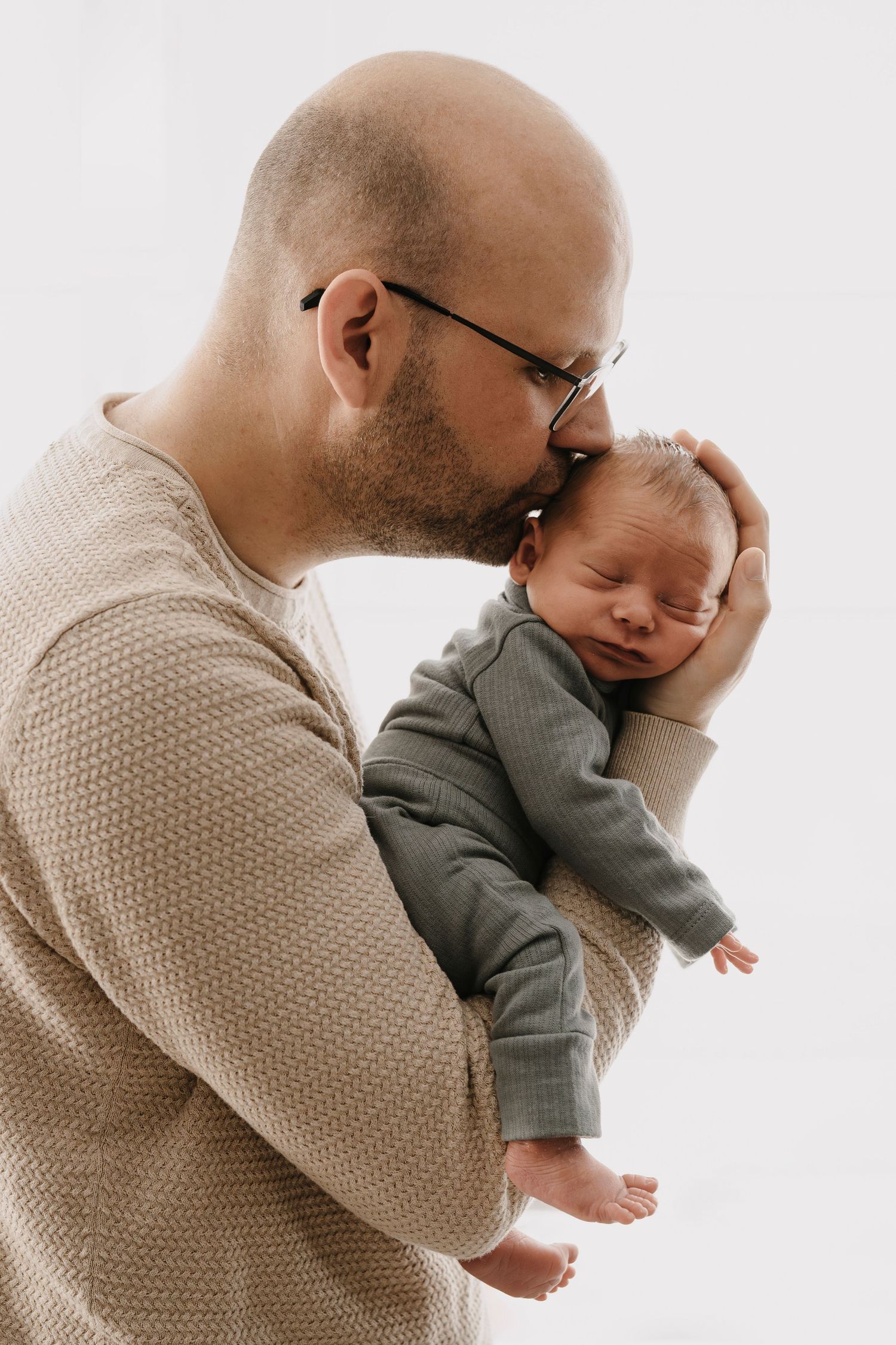 Adult in beige sweater tenderly embraces and kisses sleeping infant wearing gray outfit against white background.