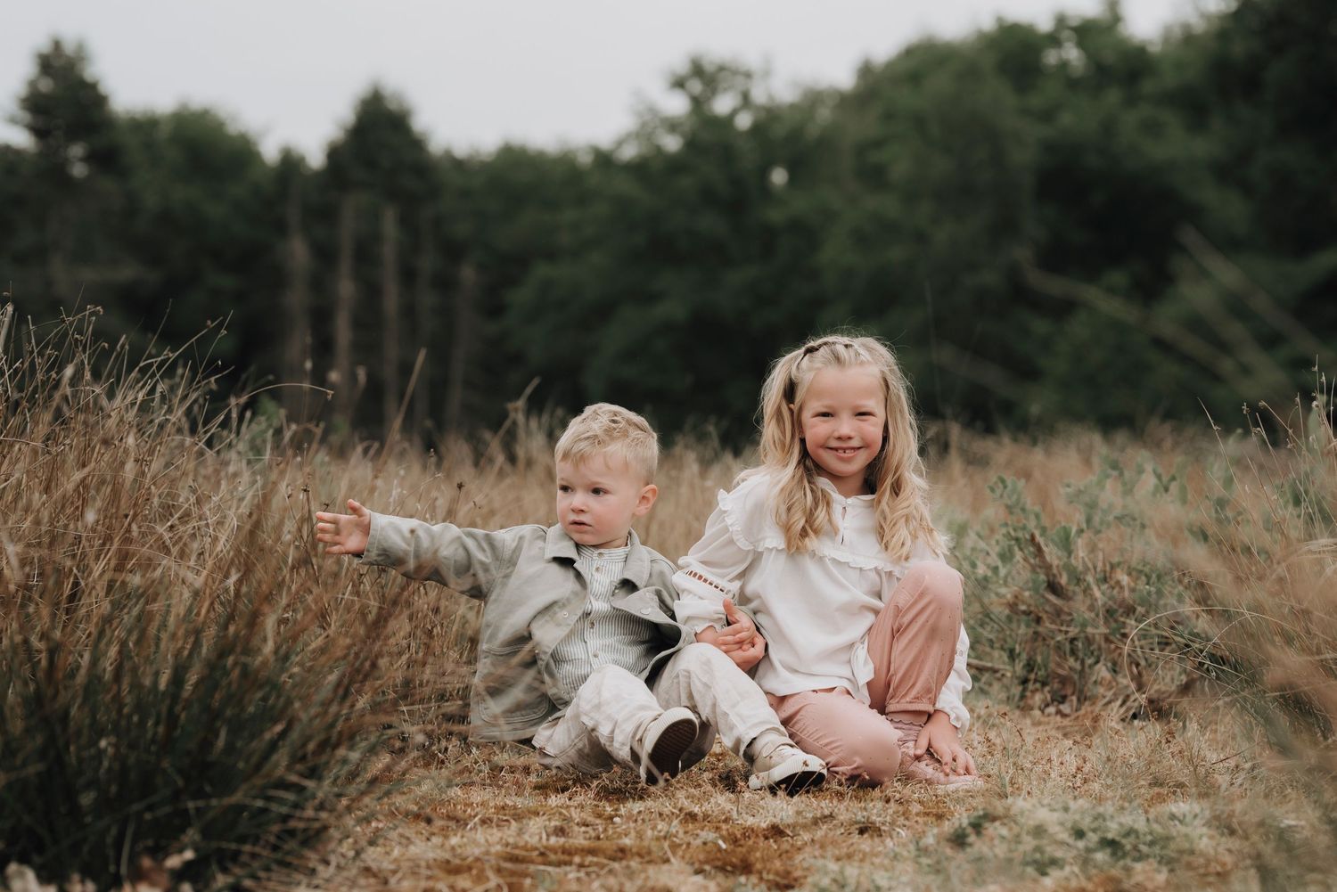 Two young children sitting in a brown field with one pointing while wearing neutral colored outfits.