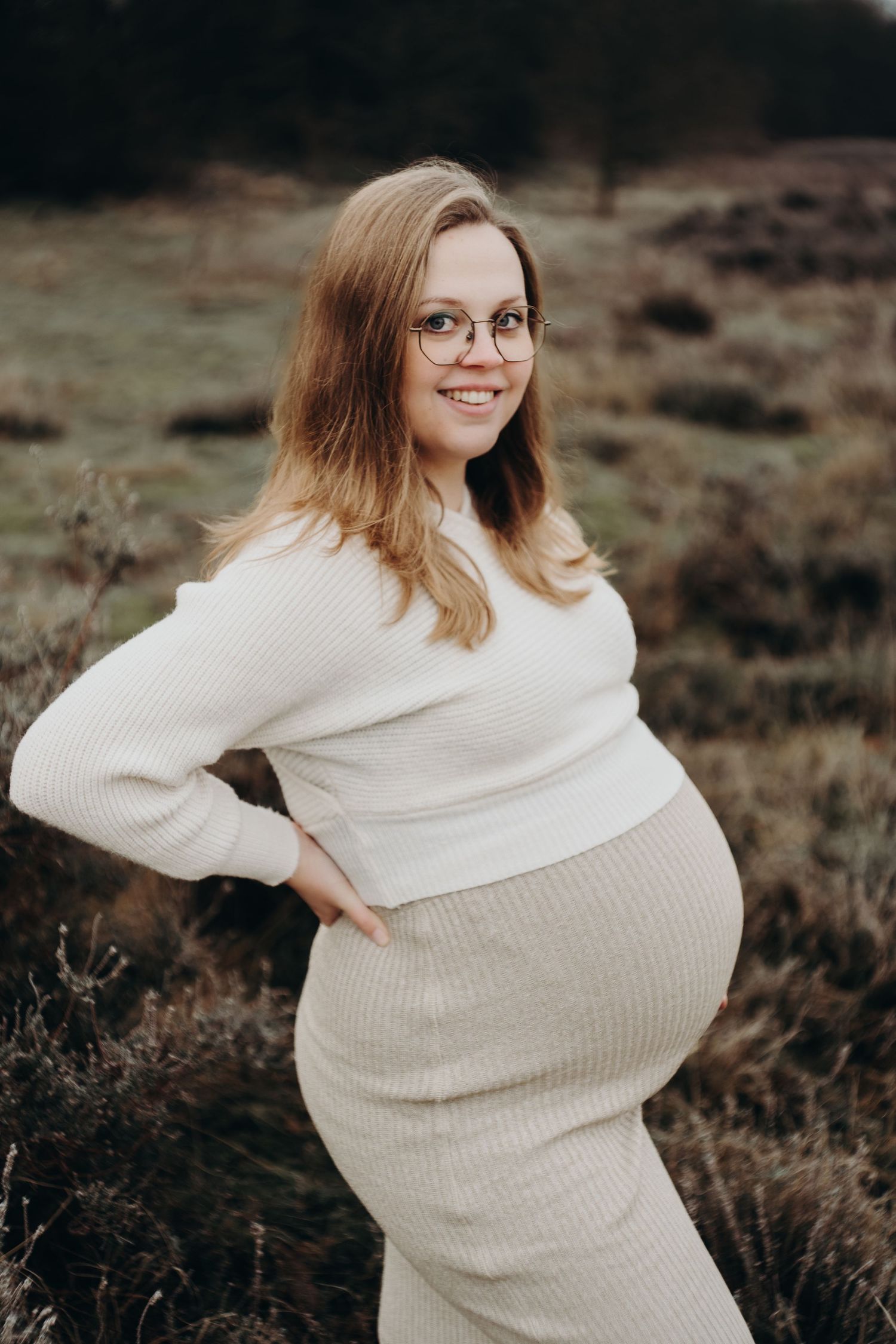 Maternity photo of woman in cream sweater dress posing in rustic outdoor setting.