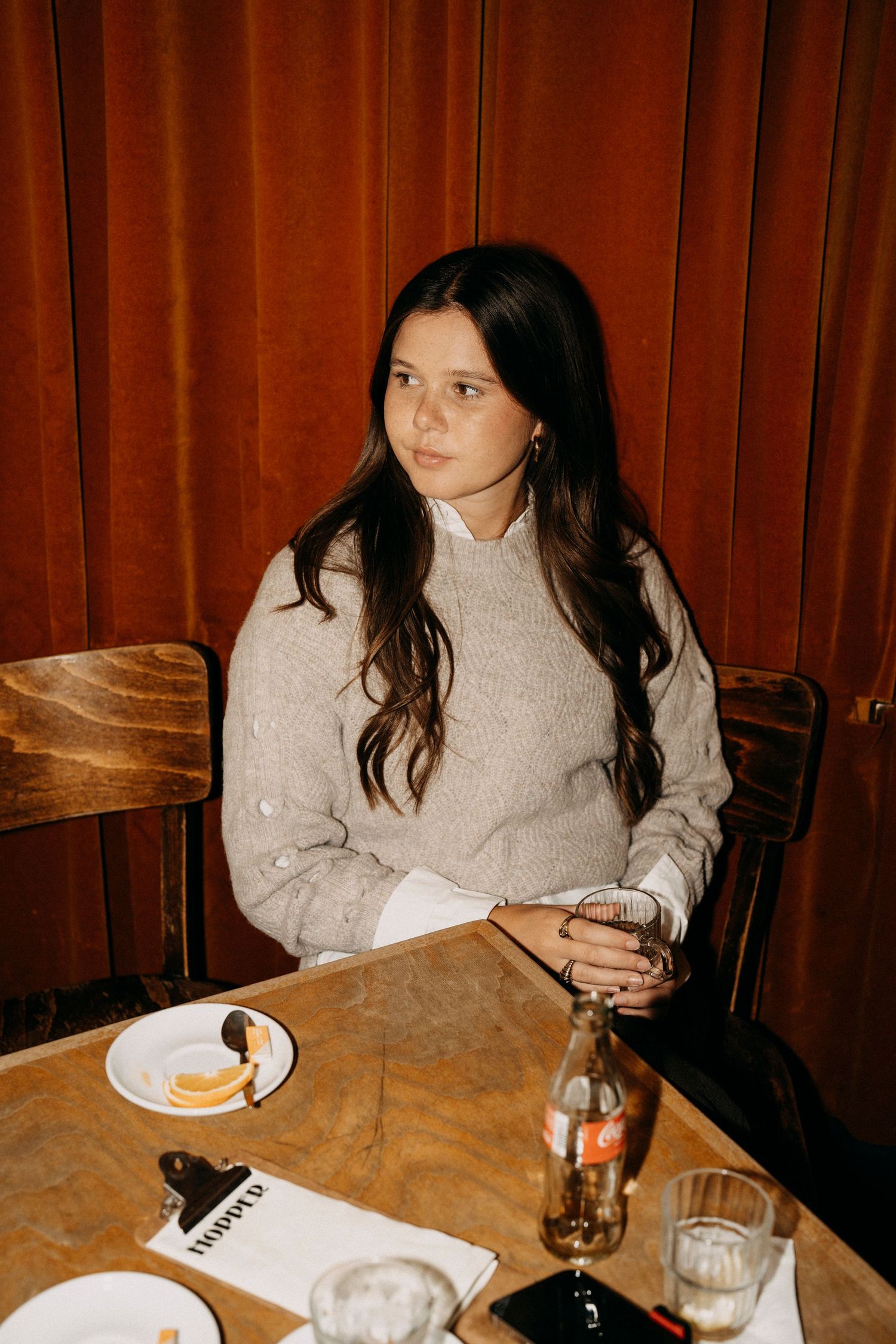 A person in a beige sweater sits at a wooden table with drinks and plates in a dimly lit restaurant setting.
