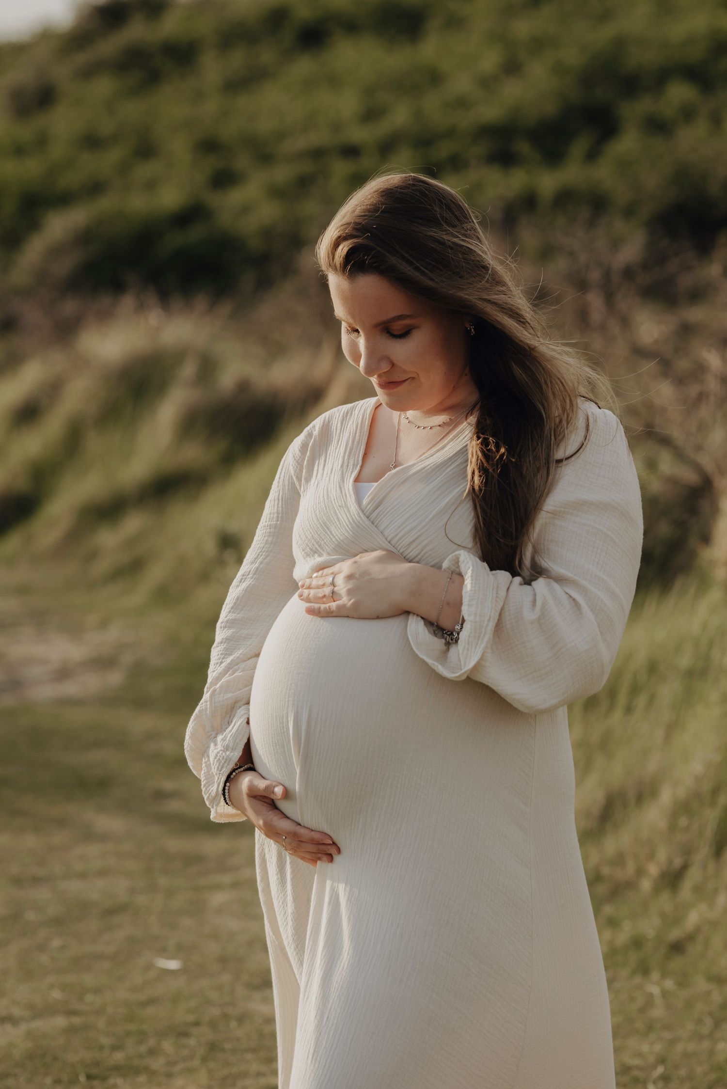 An expectant mother in a flowing white dress stands on a grassy hillside during golden hour.