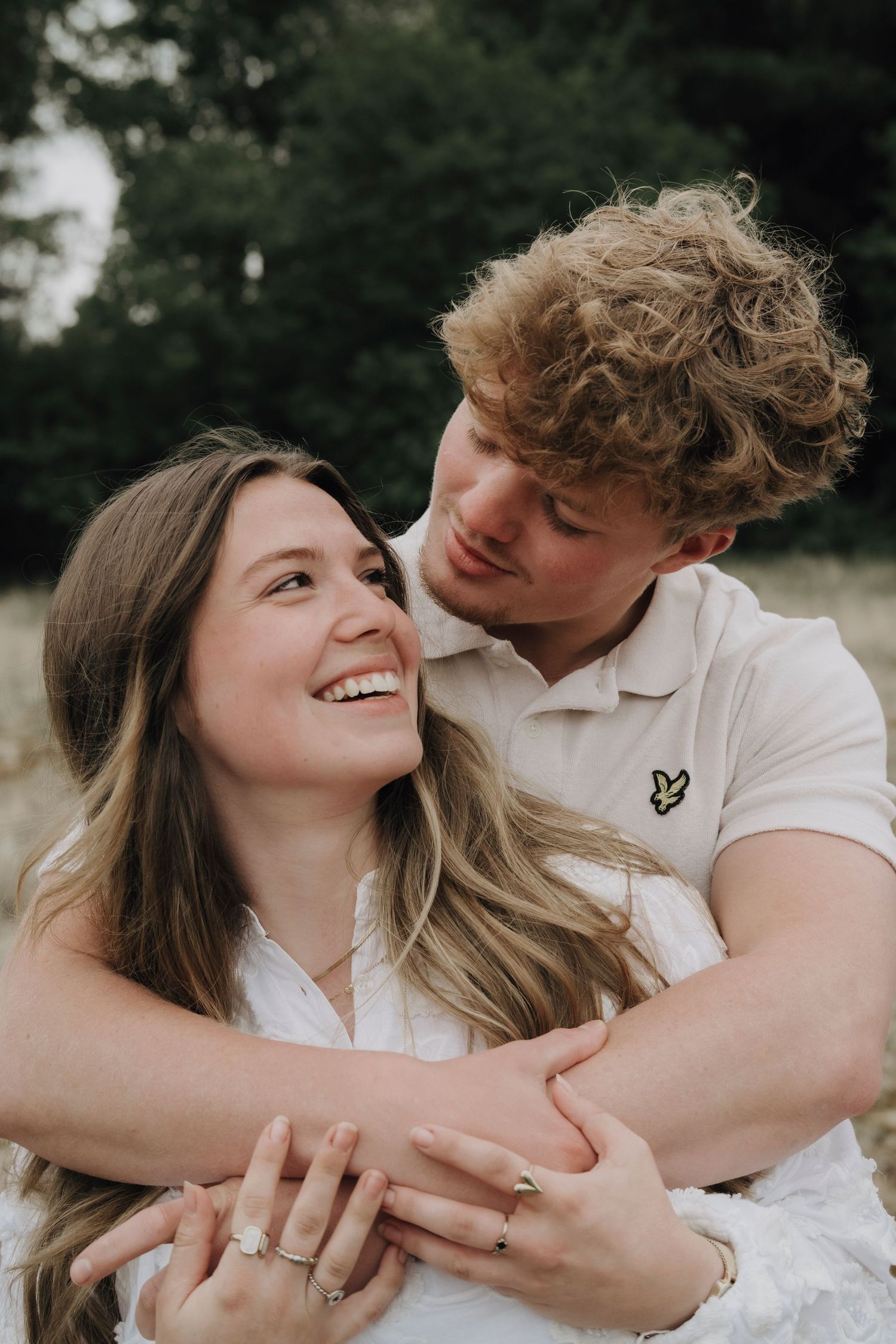 A couple embraces outdoors while sharing a joyful moment together, showing off an engagement ring.
