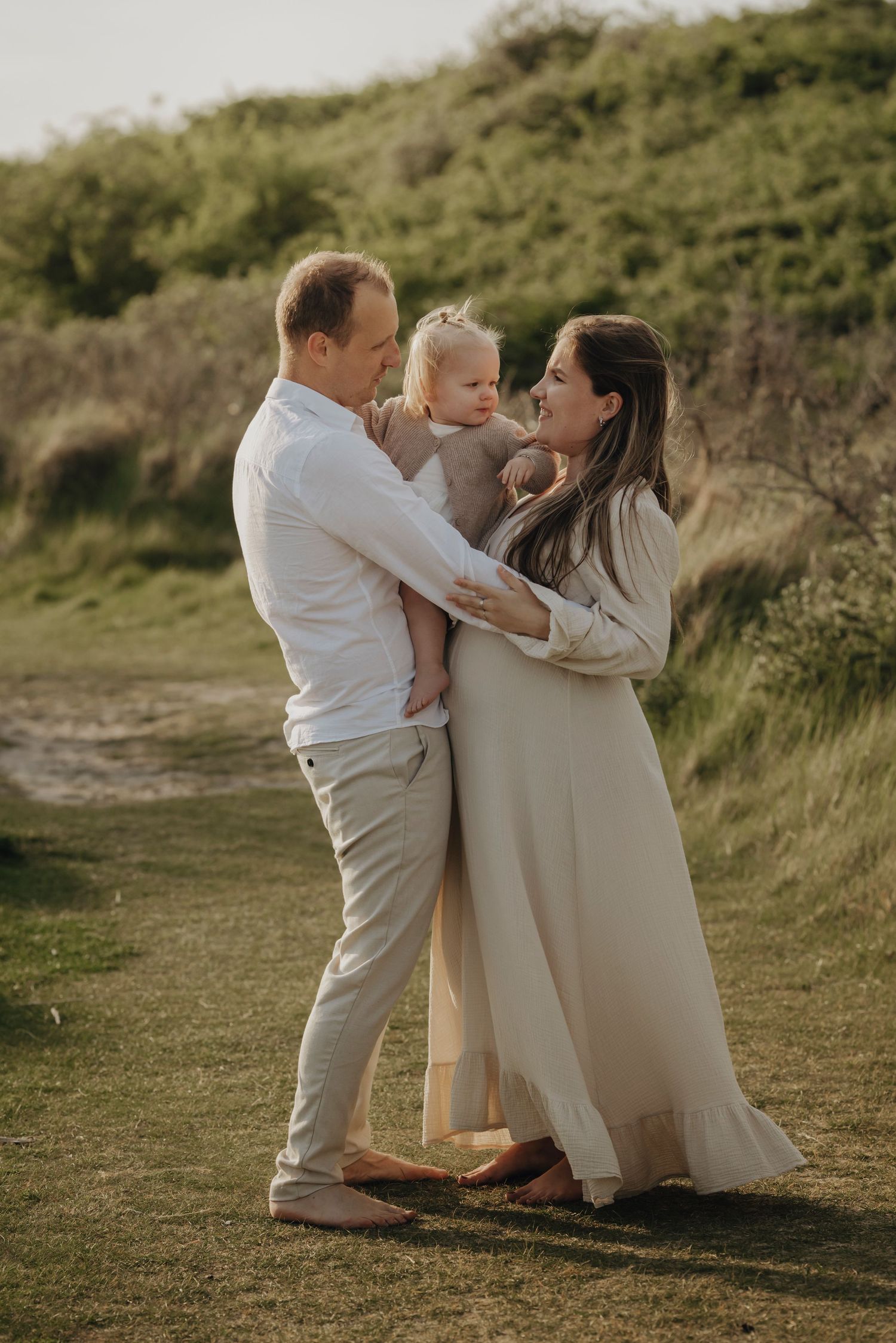Parents wearing light colored outfits share tender moment with their baby outdoors in a natural setting during sunset.