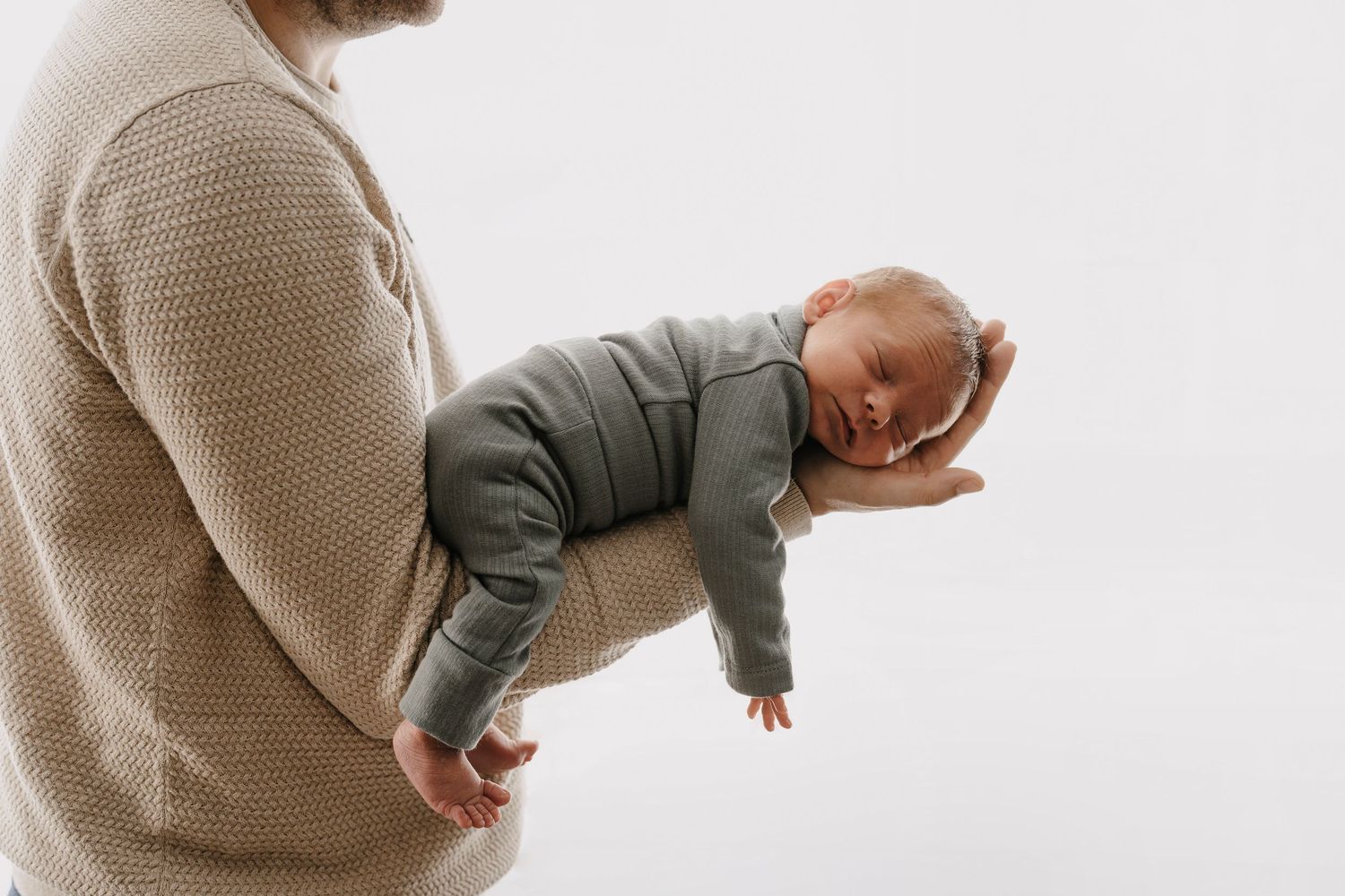 Person in beige sweater holding a sleeping baby wearing green clothing against white background.