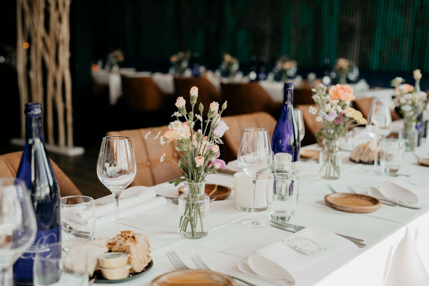 Elegant wedding reception table setting with blue wine bottles, white flowers in vases, and wine glasses on white linens.