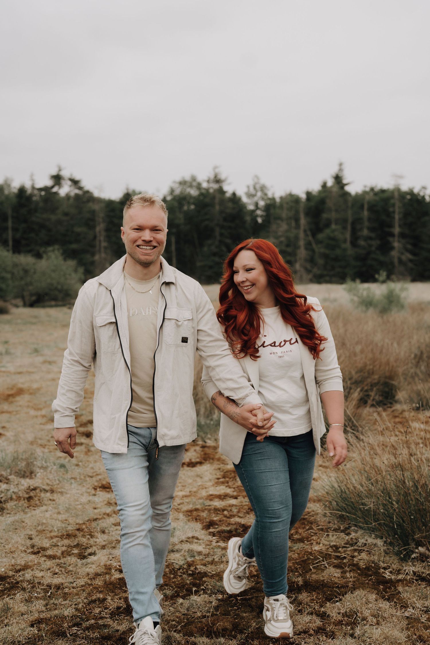 A couple walks together laughing on a nature trail with evergreen trees in the background on an overcast day.