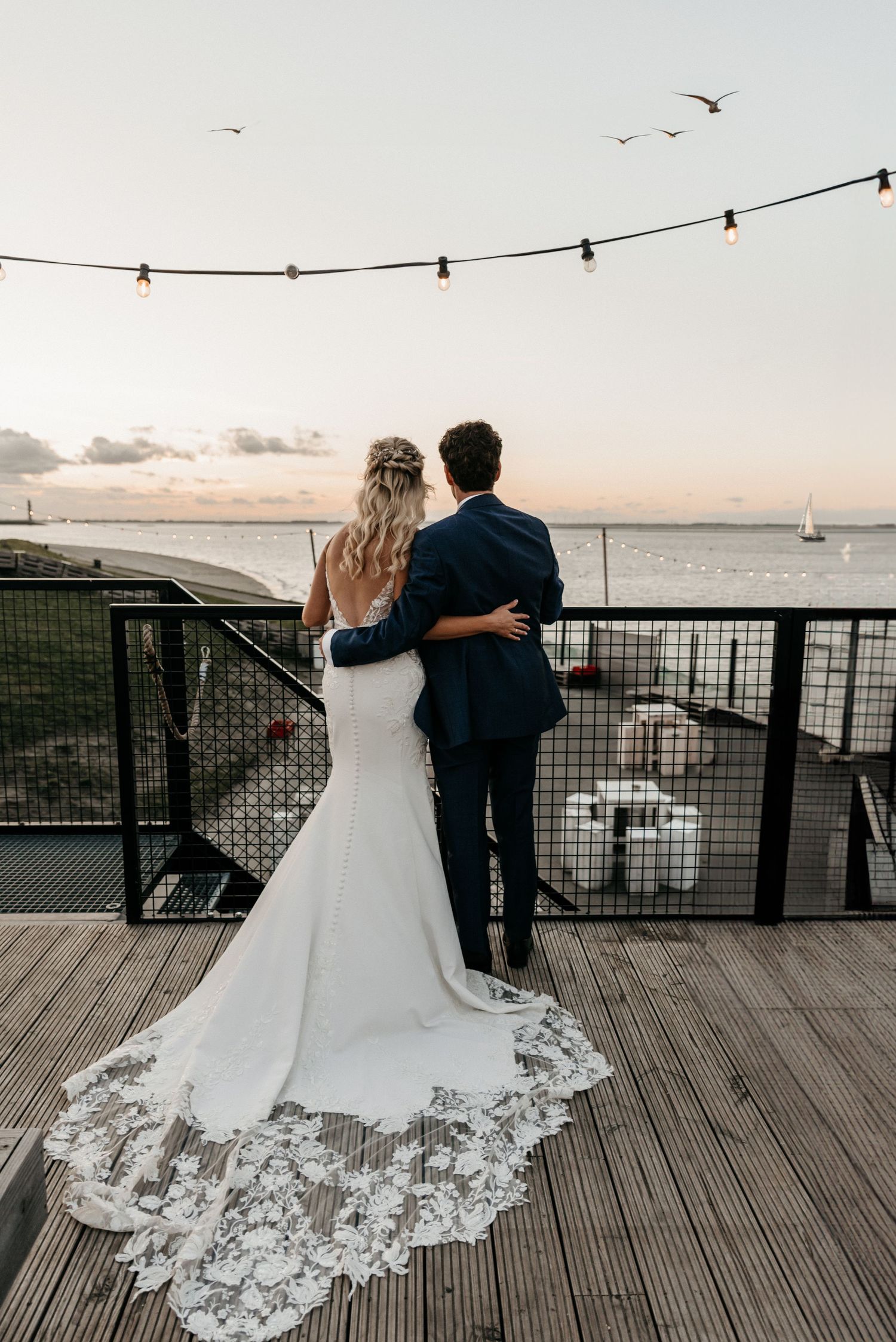 Newlyweds embrace while watching sunset, string lights overhead and lace train flowing.
