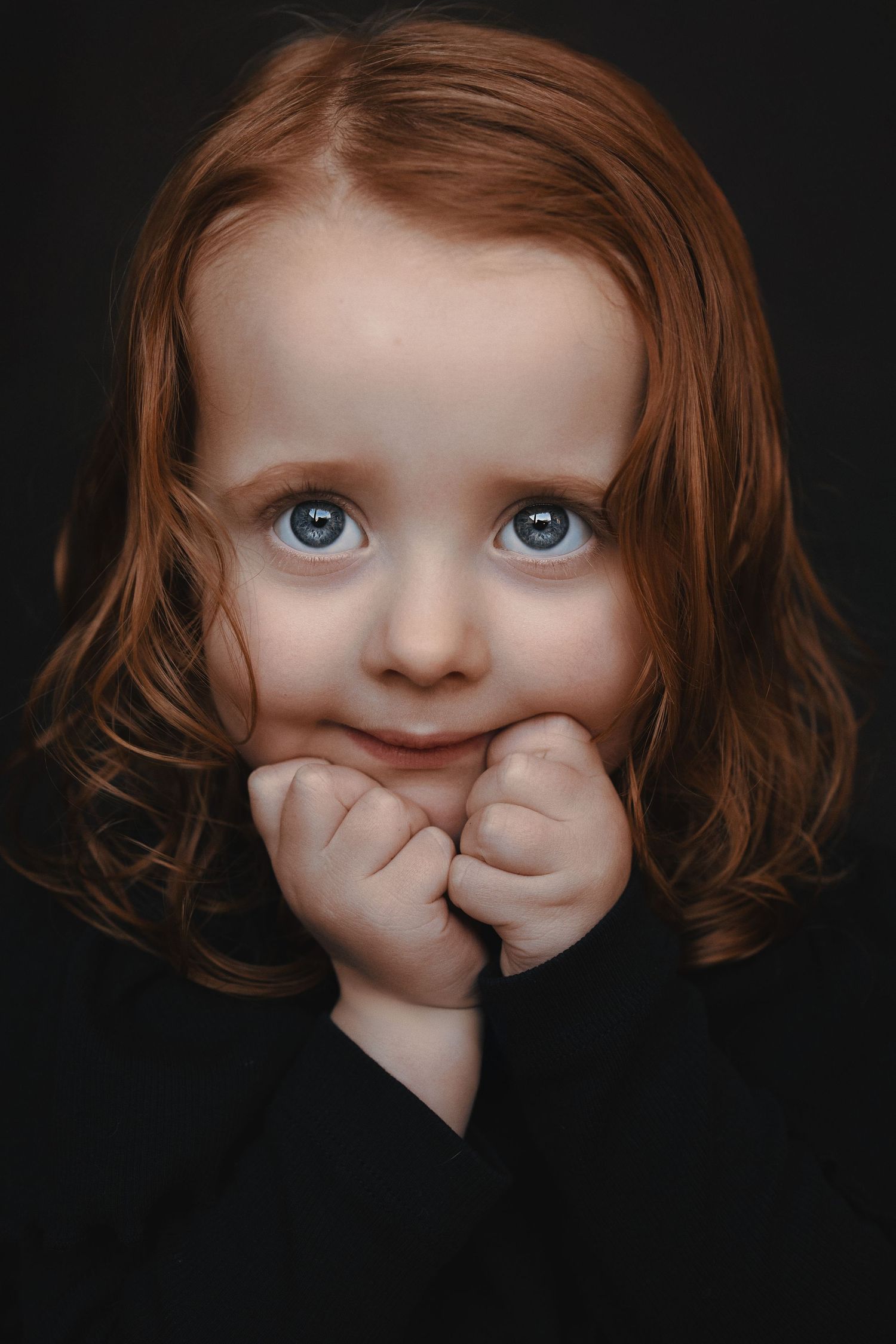 Portrait of young child with auburn curls and bright eyes against dark background.