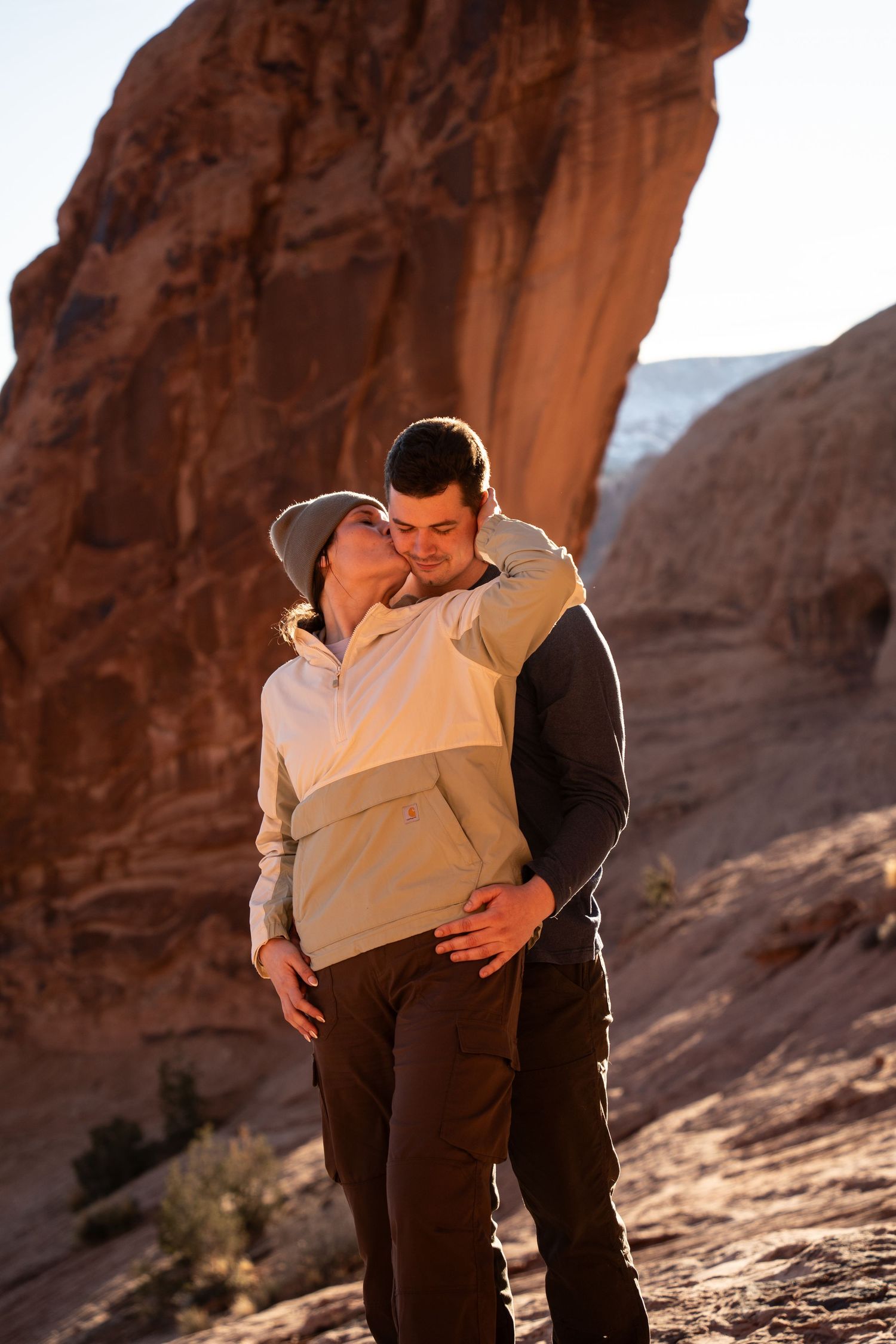 Couple sharing a romantic kiss among red rock formations in a utah desert landscape, wearing casual outdoor clothing.