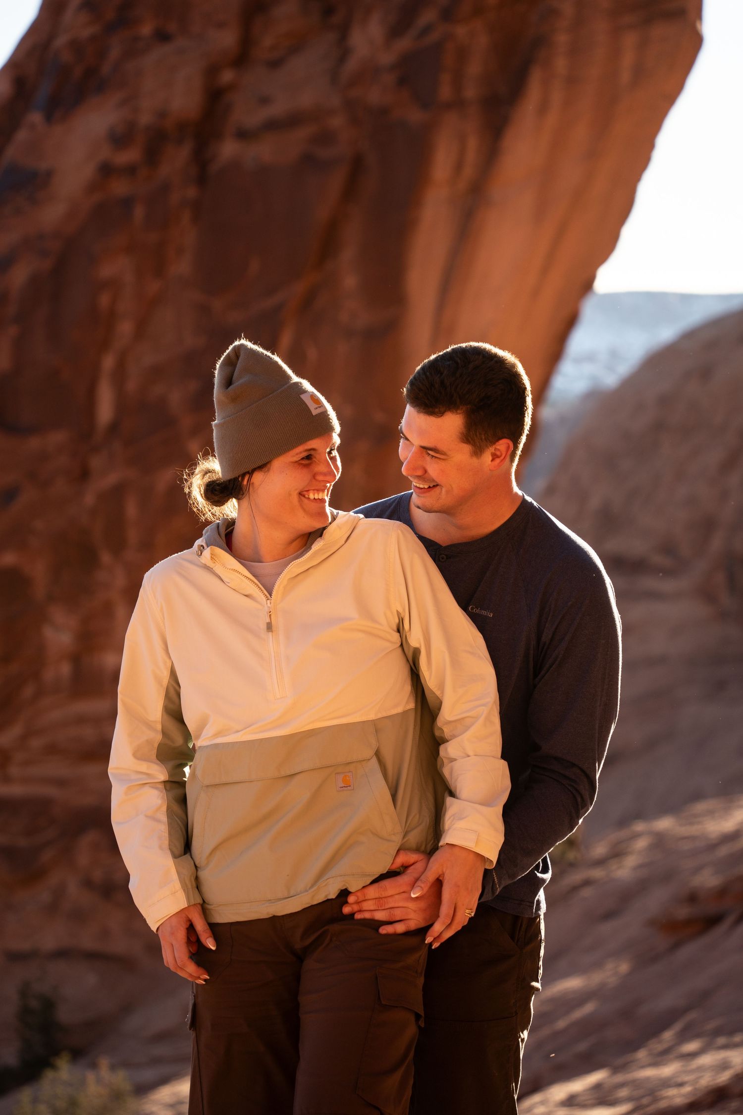 Couple smiling at each other in front of red rock formations, wearing outdoor gear in warm golden light after proposal