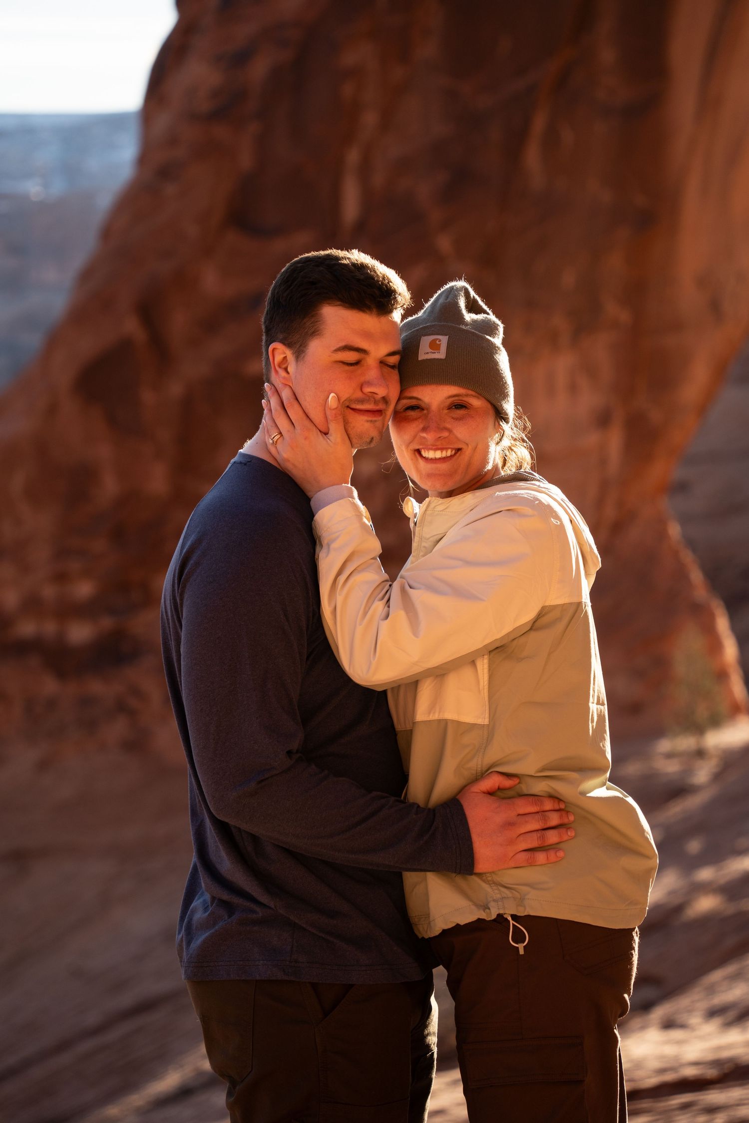 Couple embracing and smiling at sunset near red rock canyon formations corona arch moab utah, woman wearing Carhartt beanie.